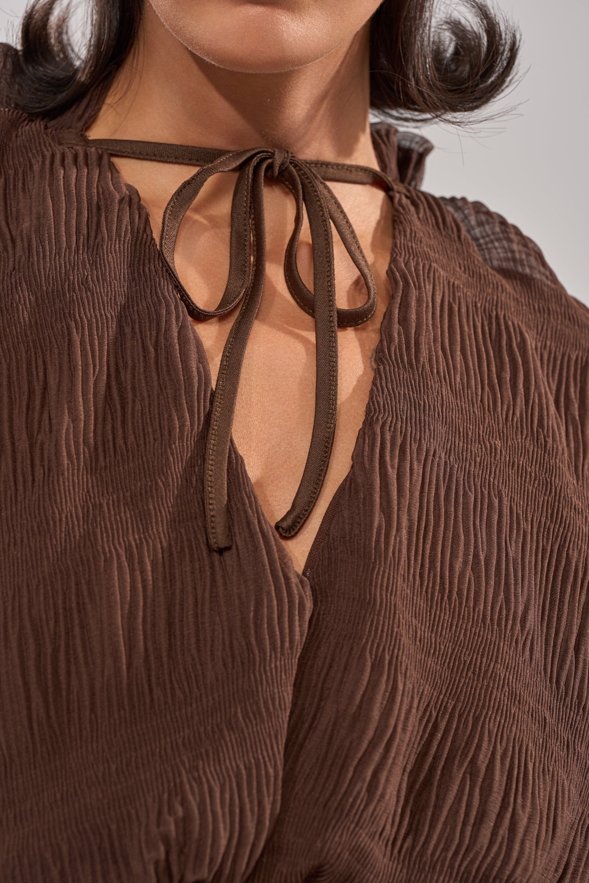 Close-up of a woman in the BABY THATS HOW I WANT IT BLOUSE—a sheer brown balloon-sleeve top with a V-neckline and thin brown tie forming a bow. Her face is partly visible, and her hair falls in loose curls. 7