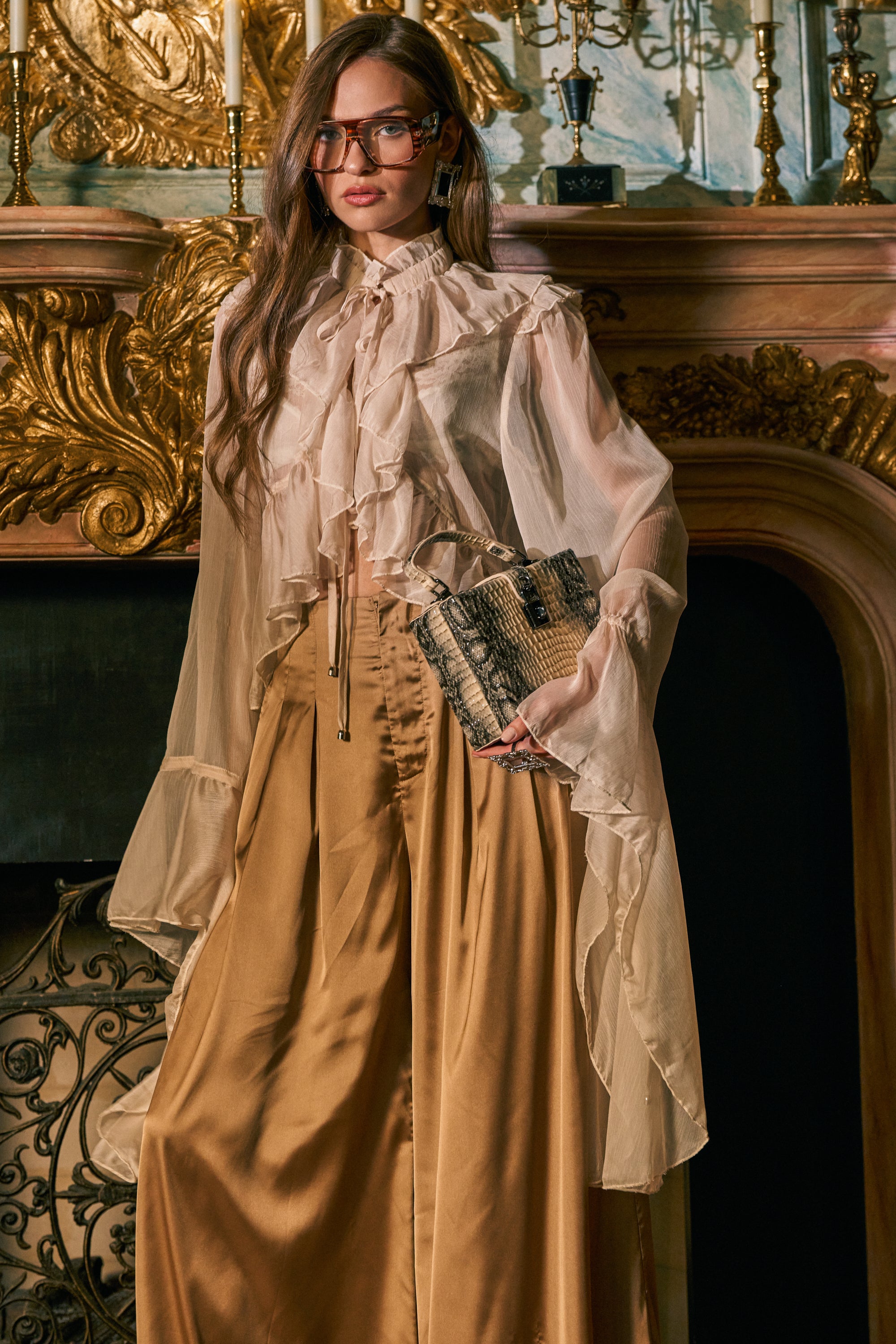 Wearing the DANCE THE NIGHT AWAY BLOUSE, a woman with wide gold satin pants stands by an ornate gold fireplace, holding a textured clutch and wearing large glasses. The romantic chiffon blouse features sheer ruffles for an elegant touch.