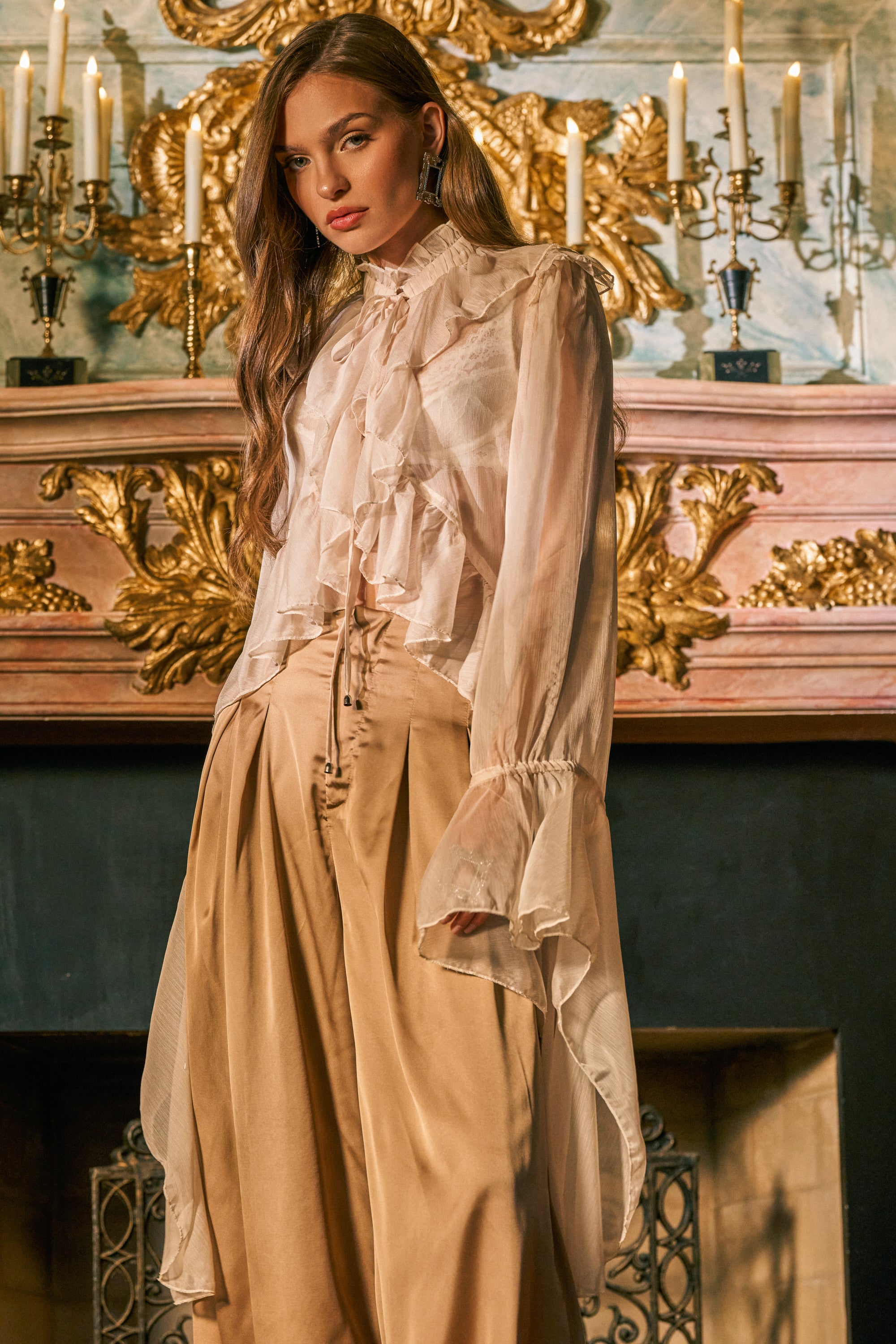 Wearing the DANCE THE NIGHT AWAY BLOUSE, a woman stands in front of an ornate fireplace adorned with gold details and glowing candles, looking at the camera.