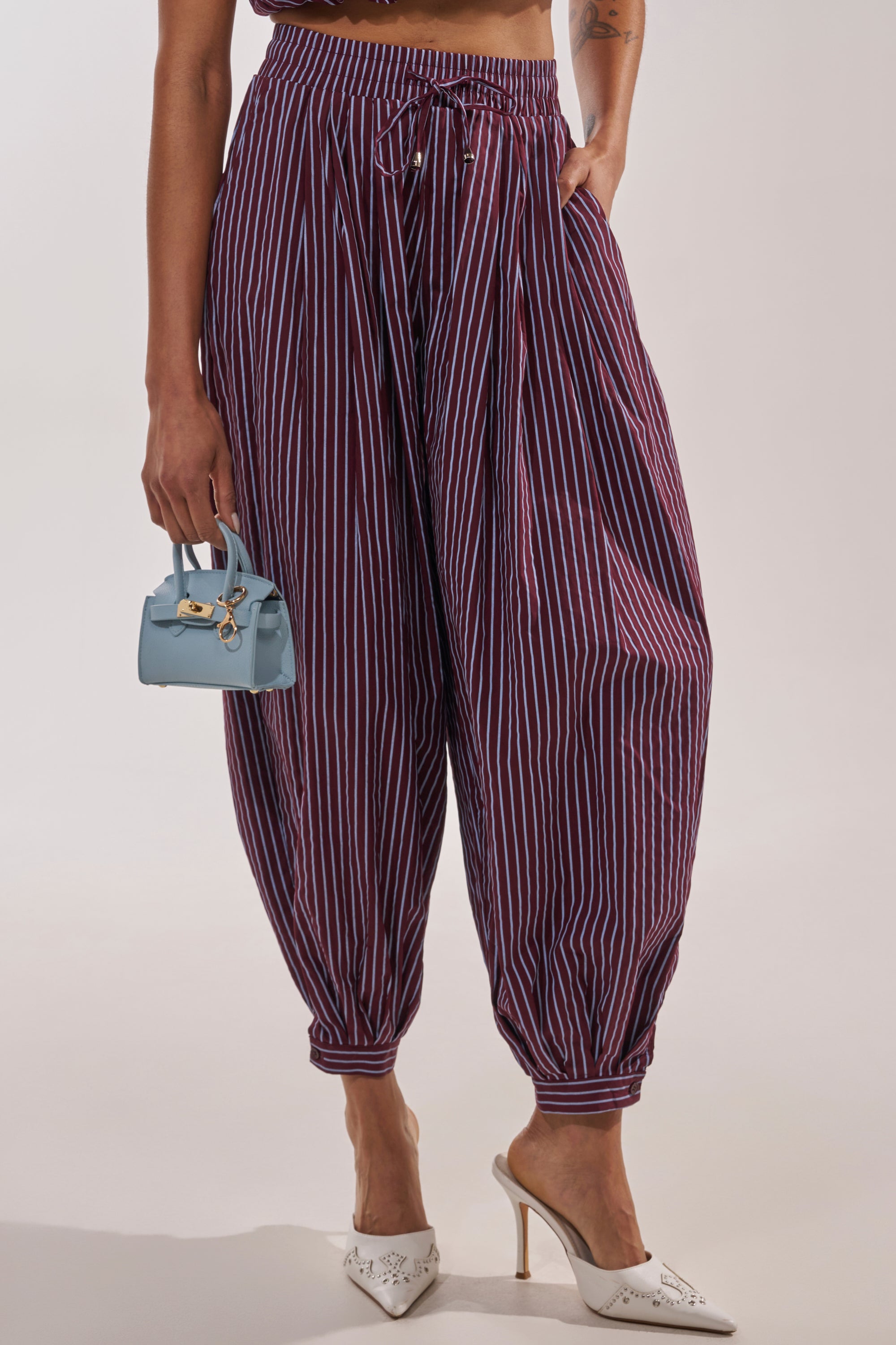 A person wearing the COULD BE DIFF BALLOON PANT in high-waisted purple with white vertical stripes, styled with white heels and a small light blue handbag, stands against a plain background. 0
