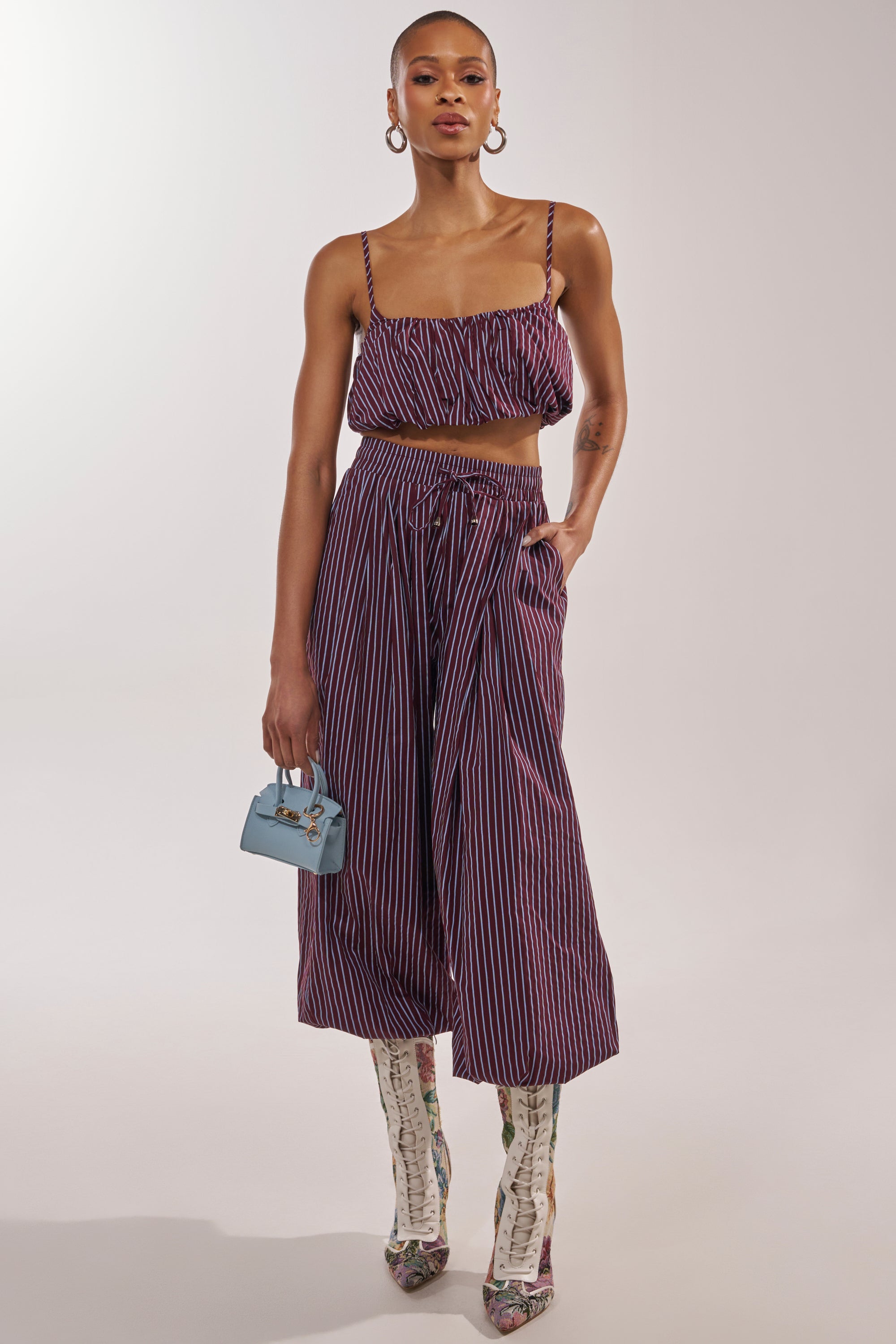 A stylish person wears purple and pink striped crop top with COULD BE DIFF BALLOON PANT in maroon and blue stripes, a small light blue handbag, hoop earrings, and knee-high patterned boots. 1