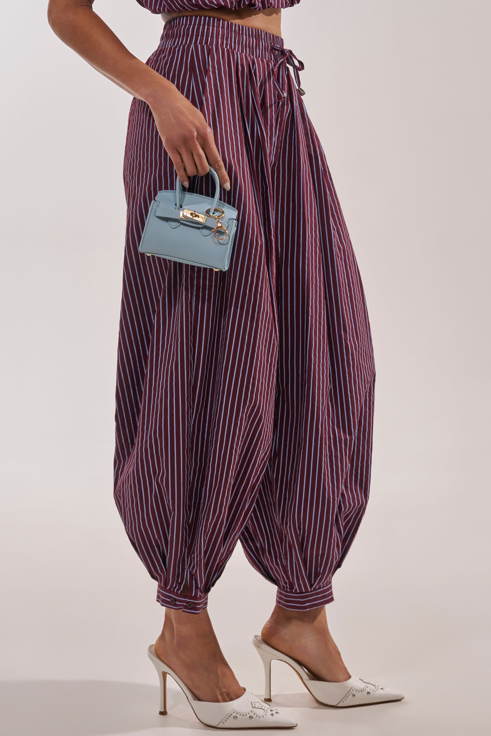Wearing the COULD BE DIFF BALLOON PANT in purple and white stripes, paired with white heels and a light blue mini handbag, the relaxed balloon-leg silhouette stands out against a plain, light background. 2