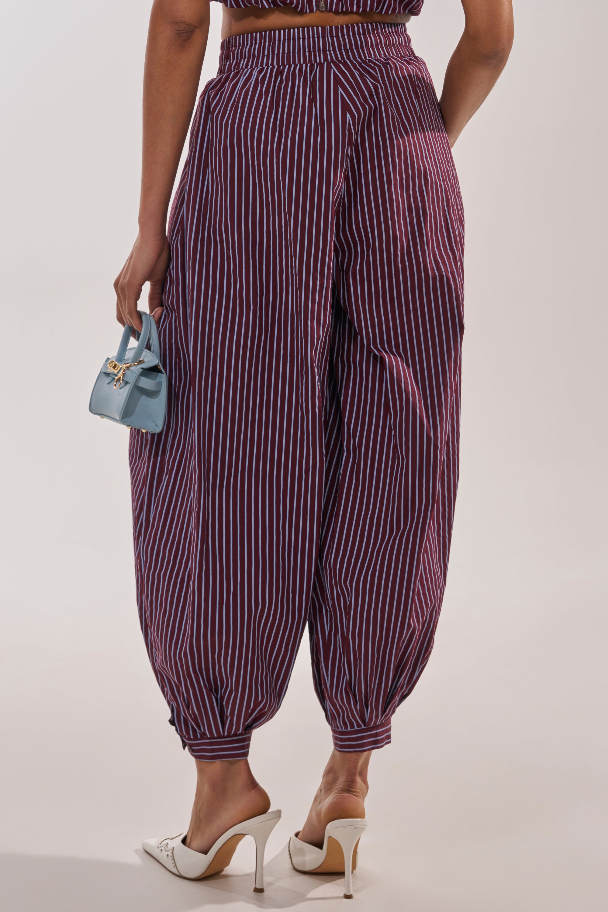 A person is seen from behind wearing COULD BE DIFF BALLOON PANT in maroon and blue stripes, paired with white high heels and holding a small light blue handbag, set against a plain background. 3