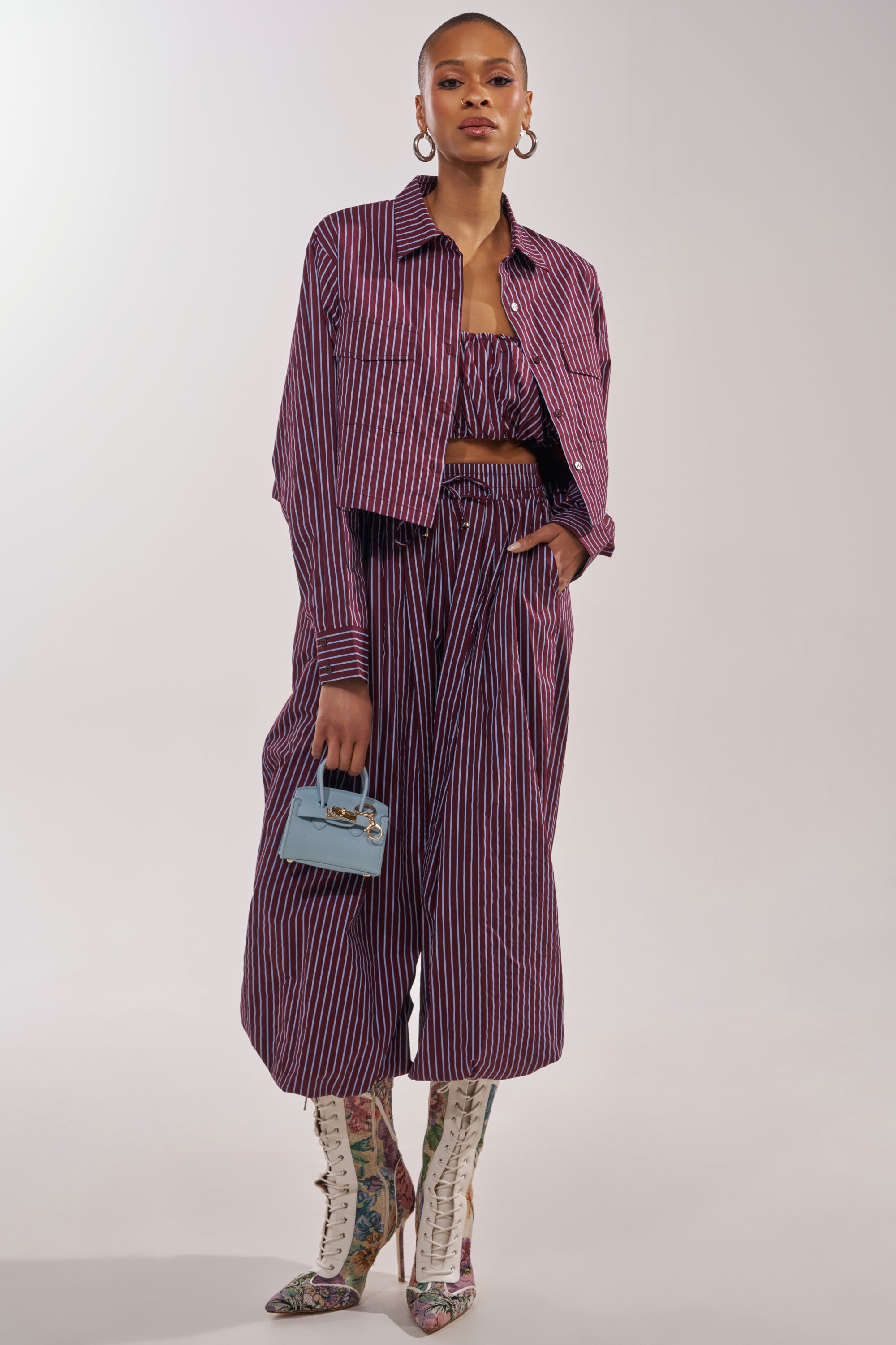 A woman wears the COULD BE DIFF BALLOON PANT as part of a purple and white striped set with a cropped jacket and strapless top. She accessorizes with a small blue handbag, lace-up floral boots, and hoop earrings. 5