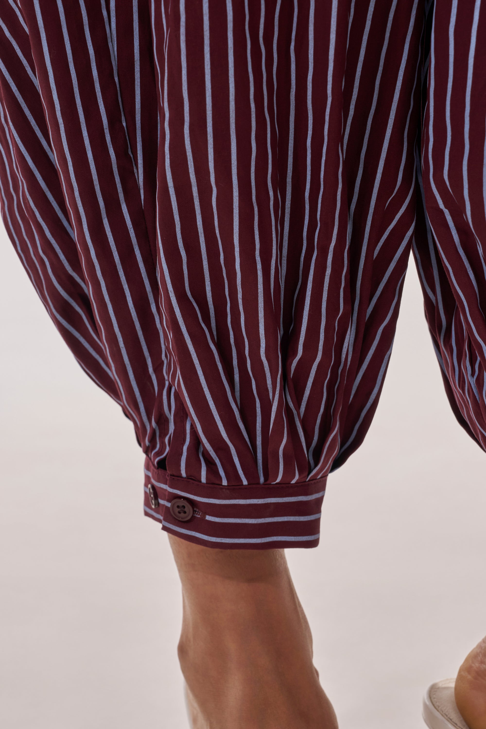 Close-up of a person’s lower legs wearing the COULD BE DIFF BALLOON PANT in maroon and blue stripes. The relaxed balloon-leg silhouette features cuffed ankles with button closures. The person is barefoot on a neutral background. 7