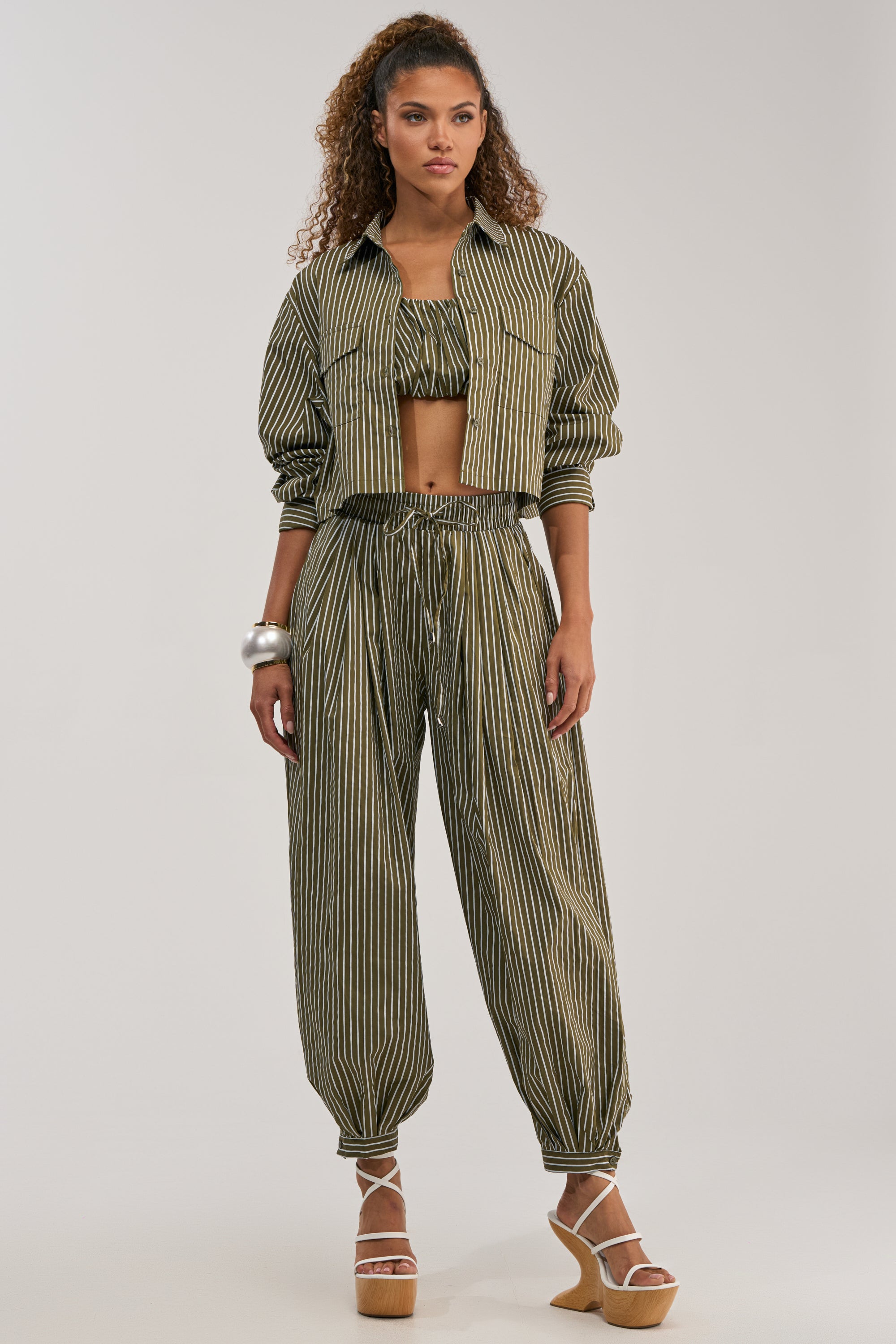 Wearing the COULD BE DIFF BALLOON PANT, a woman stands against a plain background in stylish summer fashion—her green and white striped crop jacket, matching smocked tube top, and elastic-cuff pants paired with platform sandals and silver bangles create an effortlessly chic look.