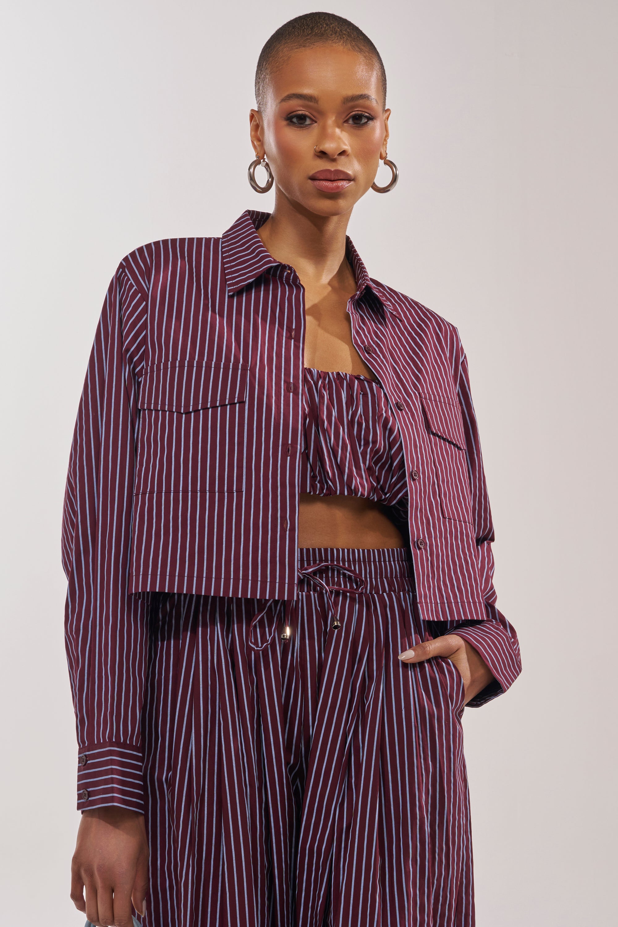 A woman with a shaved head rocks a smart-casual look, pairing the COULD BE DIFF BUTTON DOWN in maroon and blue stripes over a ruffled crop top and high-waisted pants. Large hoop earrings complete her stylish ensemble. 0