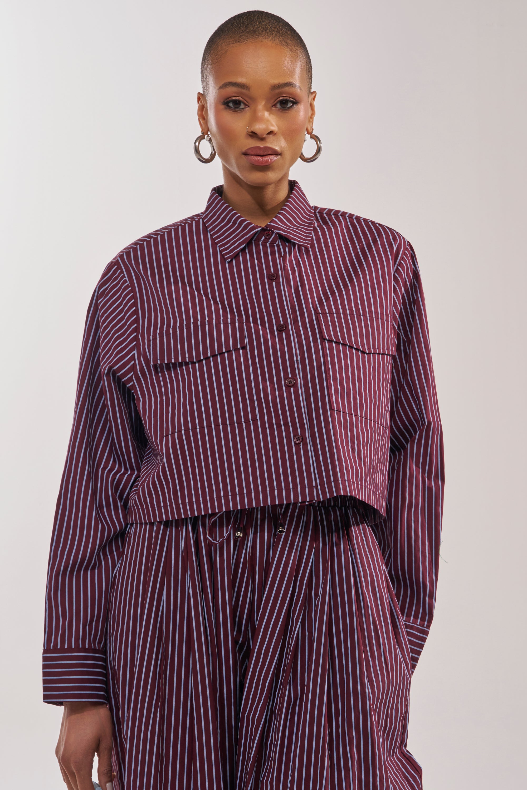 A woman with short hair wears the COULD BE DIFF BUTTON DOWN—a maroon and blue striped cropped shirt—styled smart-casual with matching wide-leg pants and hoop earrings, confidently facing the camera against a plain background. 1