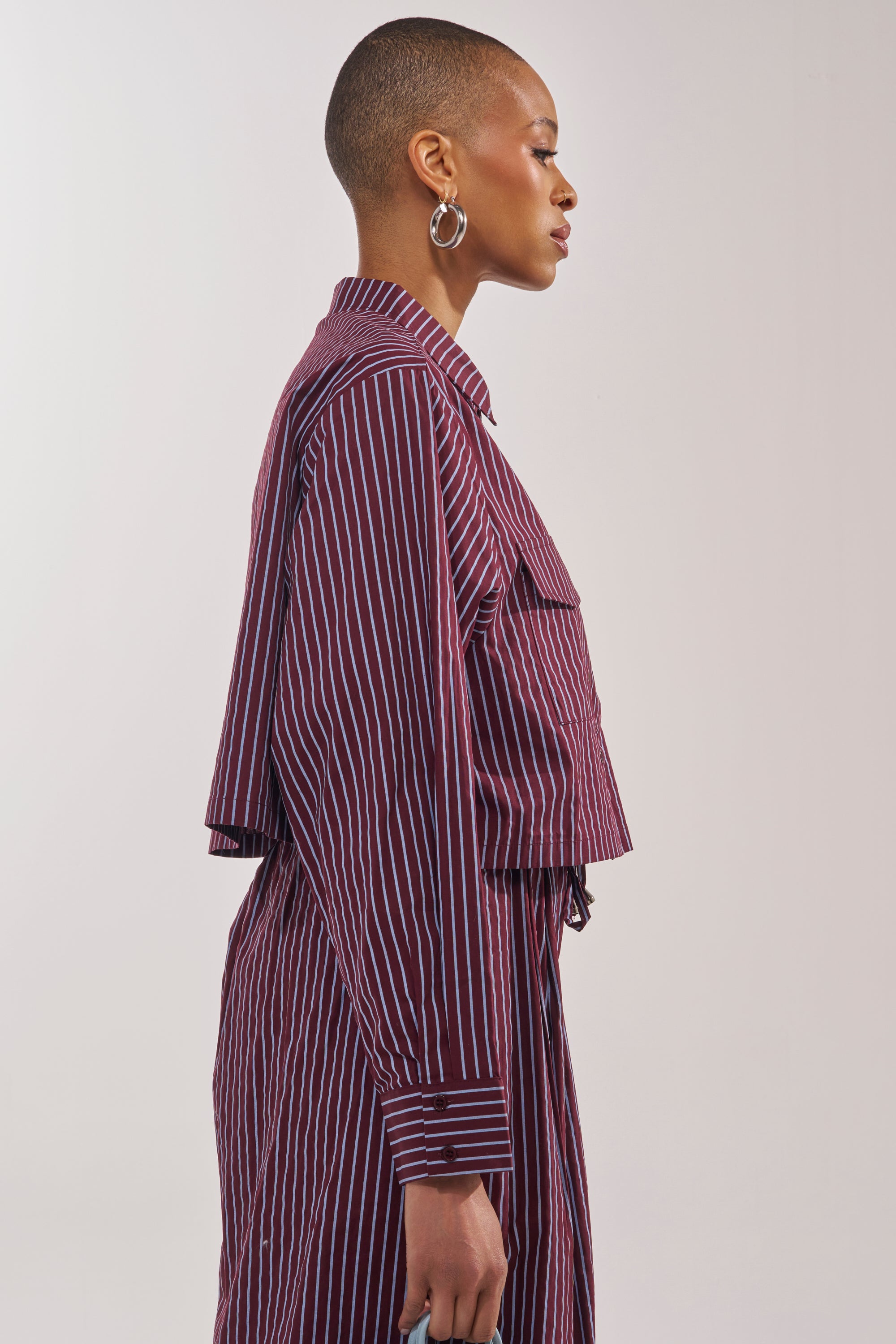 A person with a shaved head, large hoop earrings, and a maroon and blue striped COULD BE DIFF BUTTON DOWN matching set stands in profile against a plain light background. 2