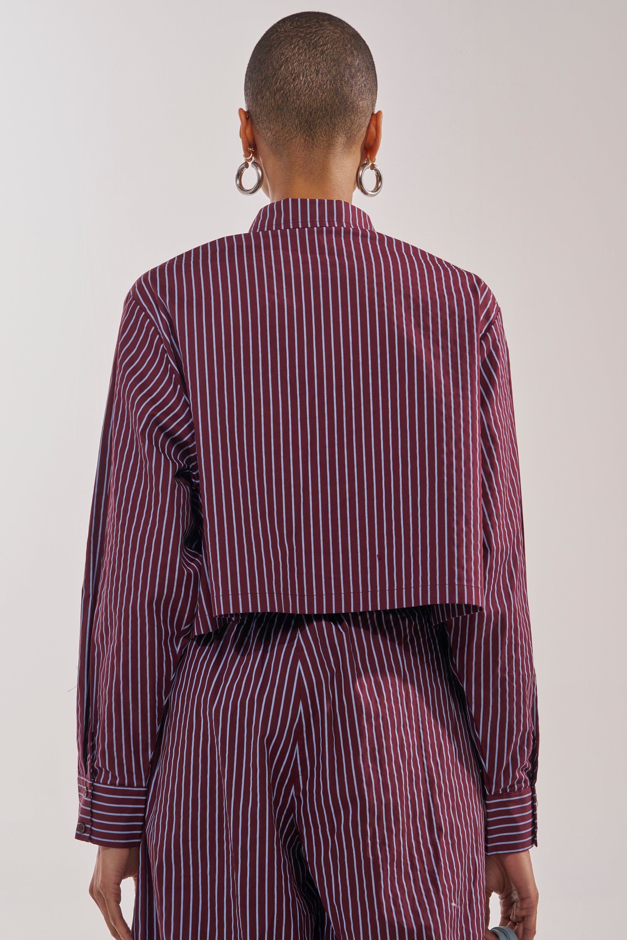 A person with a shaved head and hoop earrings is shown from behind, wearing the COULD BE DIFF BUTTON DOWN—a maroon and blue striped, long-sleeve cropped shirt with a boxy silhouette—paired with matching pants. 3