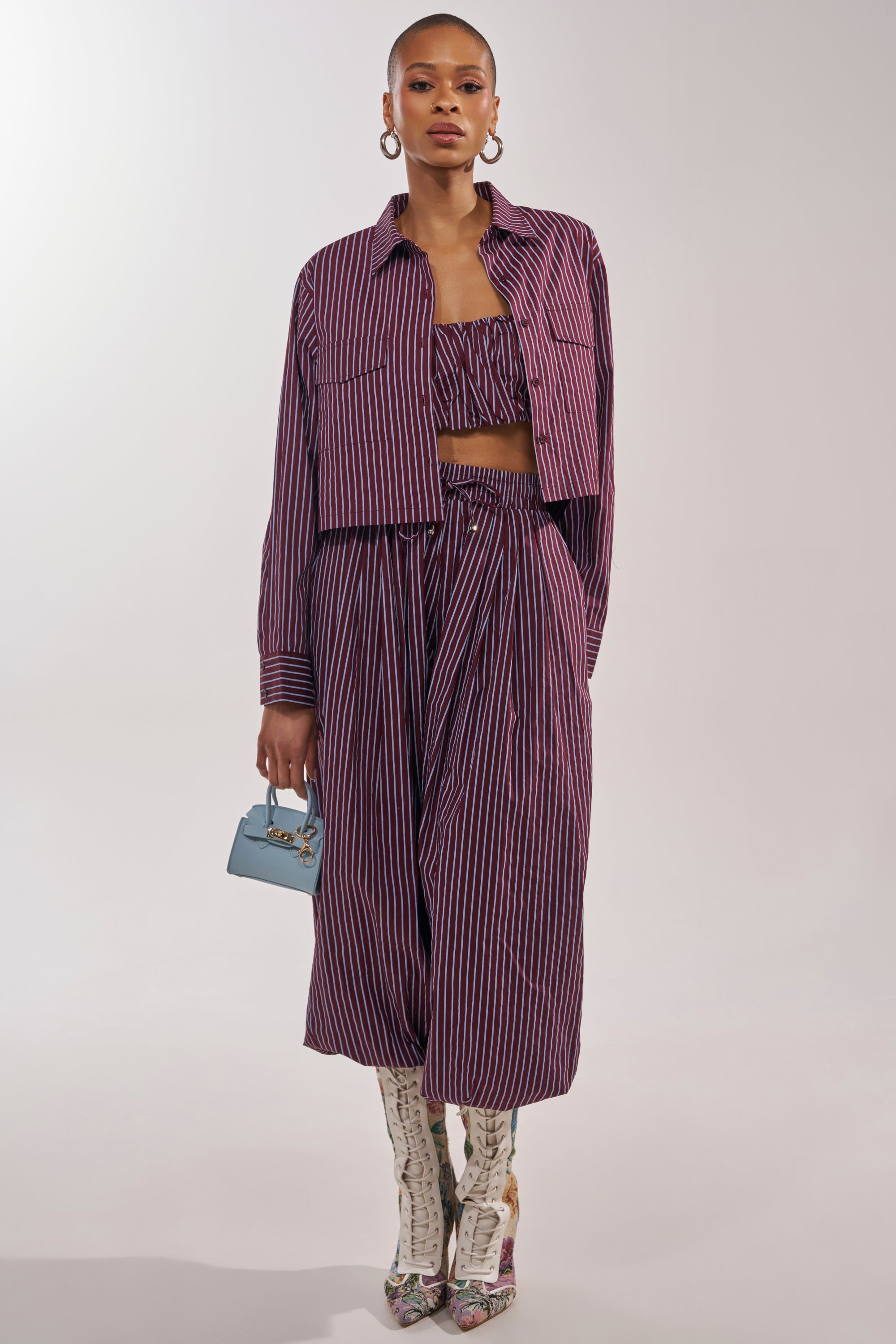 A woman models the COULD BE DIFF BUTTON DOWN, a maroon and blue striped, structured crop top, paired with a matching jacket and draped skirt. She completes the look with hoop earrings, a blue handbag, and patterned knee-high boots. 5