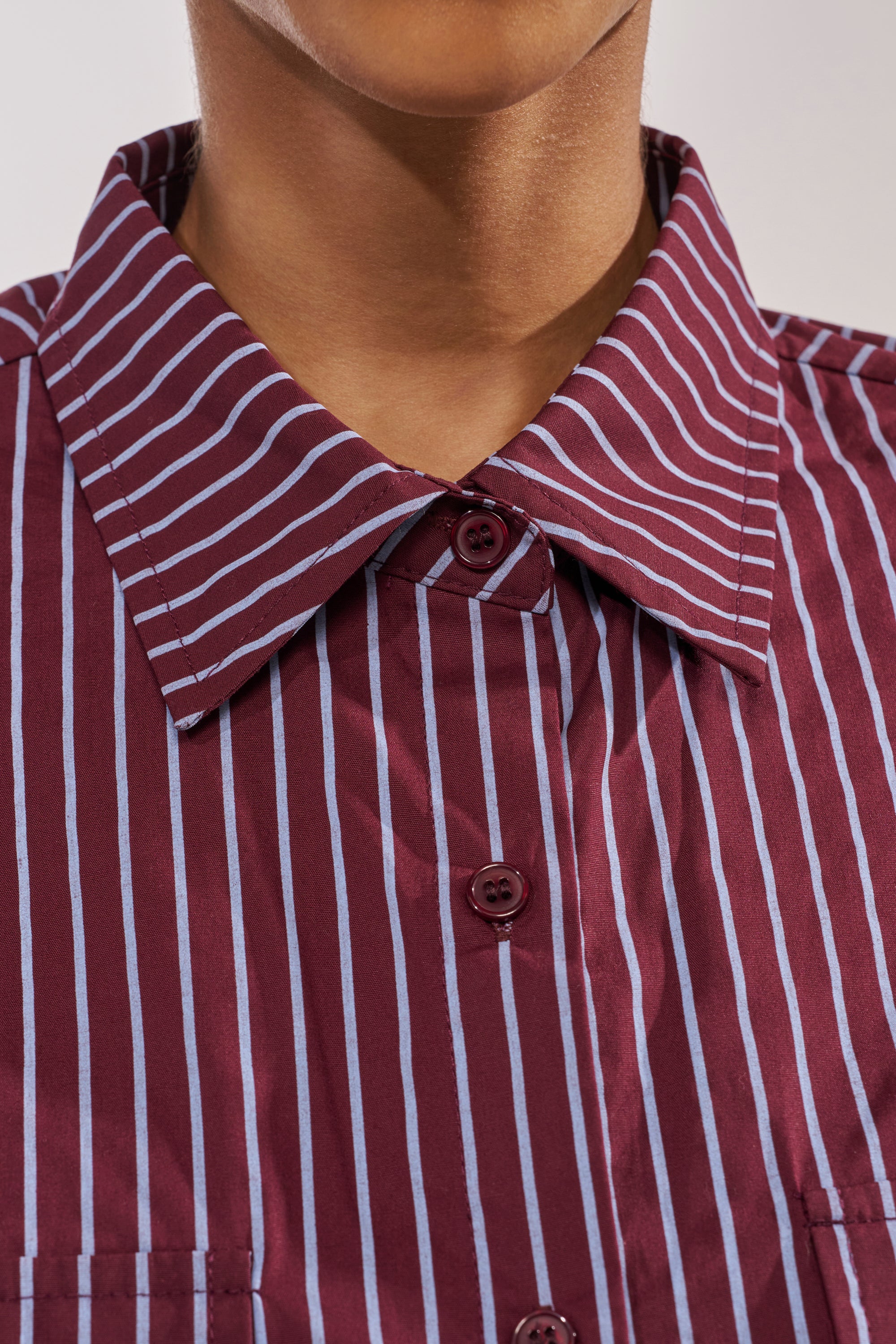 Close-up of a person wearing the COULD BE DIFF BUTTON DOWN, a maroon and blue striped cropped shirt with a pointed collar. The shirt is buttoned to the top, showing only the lower part of their face and neck. 6