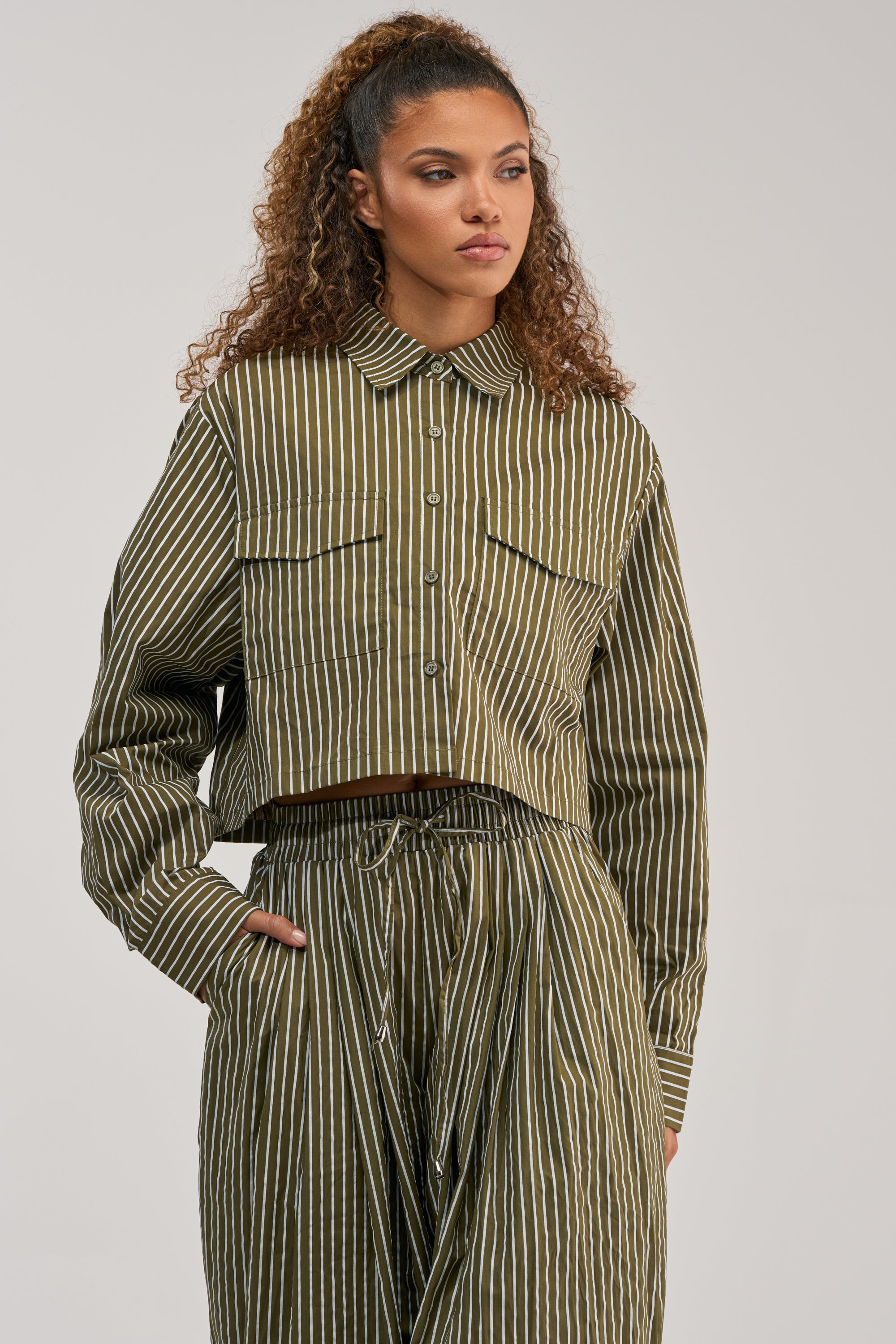 A woman stands with one hand in her pocket, looking to the side against a plain background, wearing the COULD BE DIFF BUTTON DOWN—an olive and white vertically striped blouse paired with matching pants.
