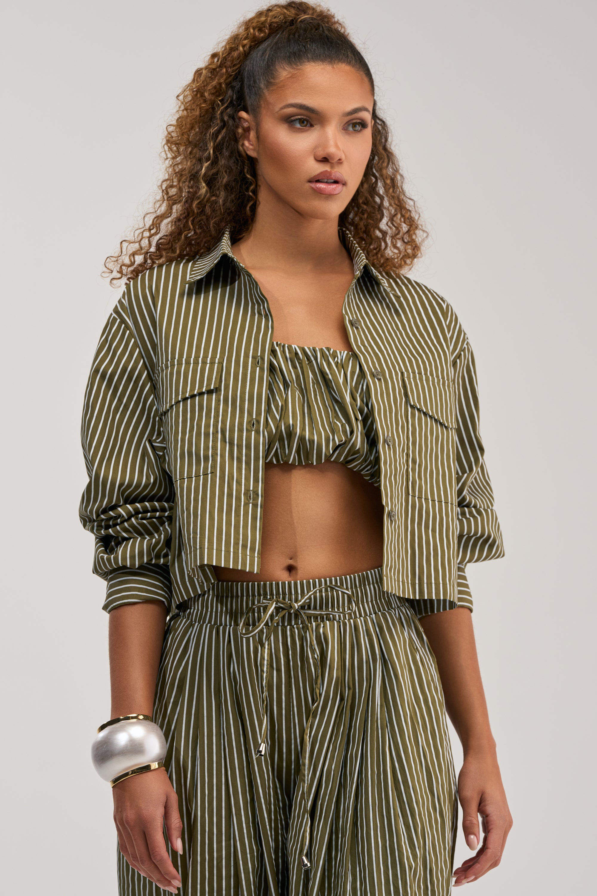 A woman stands wearing the COULD BE DIFF BUTTON DOWN, an olive green and white vertical striped blouse styled as a cropped jacket, paired with a matching bralette and pants. Her look channels the refined edge of this striped button-down, adding a chic twist to business casual. She has long, curly hair in a high ponytail and accessorizes with a large silver bangle on her left wrist.