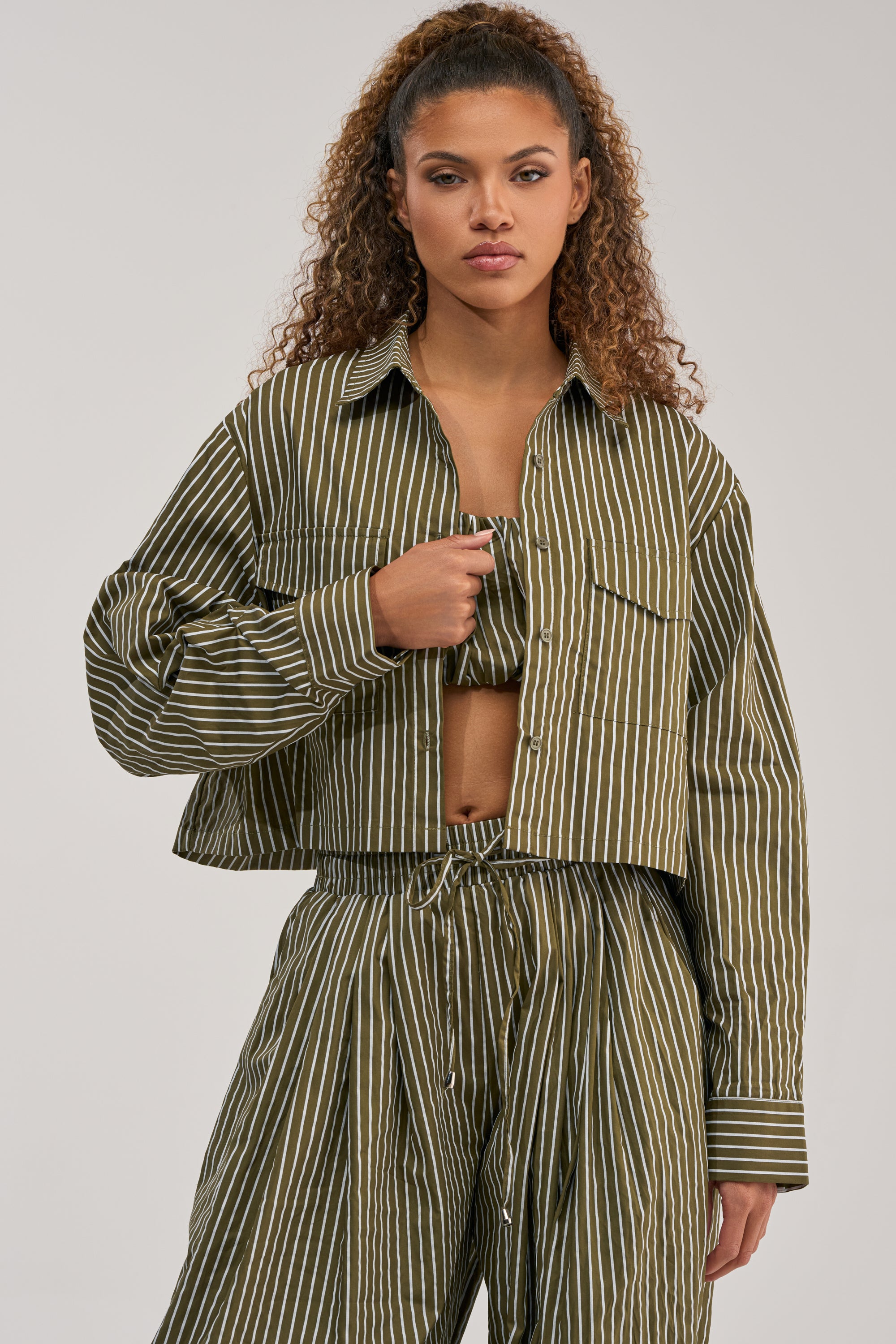 Wearing the COULD BE DIFF BUTTON DOWN, a woman with curly hair stands against a plain background in a matching olive striped blouse and pants set, completed with a coordinating jacket.