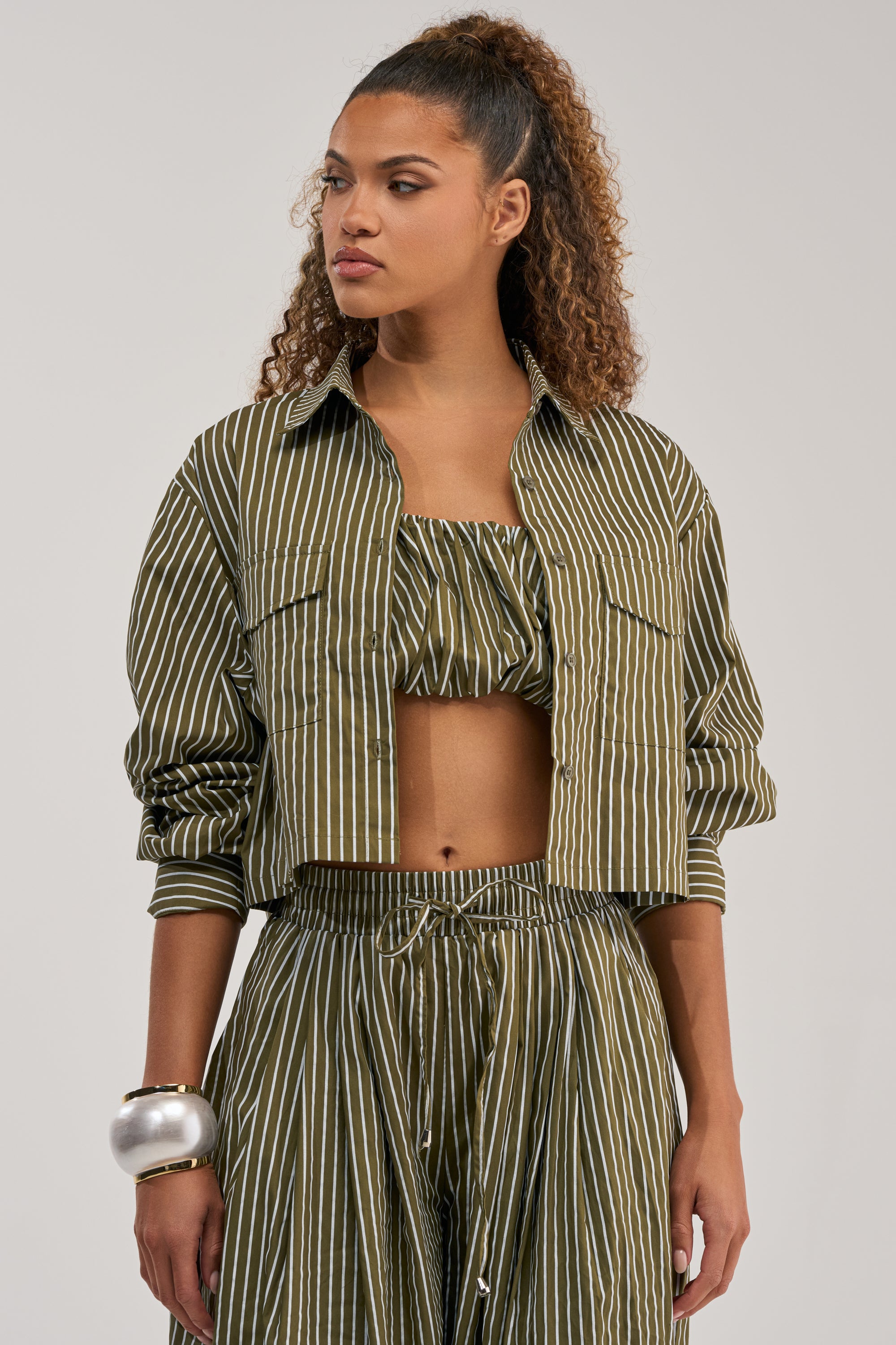 Wearing the COULD BE DIFF BUTTON DOWN blouse layered under a cropped jacket and paired with a bandeau top and wide-leg pants, she styles her curly hair in a high ponytail and accessorizes with a chunky silver bracelet.