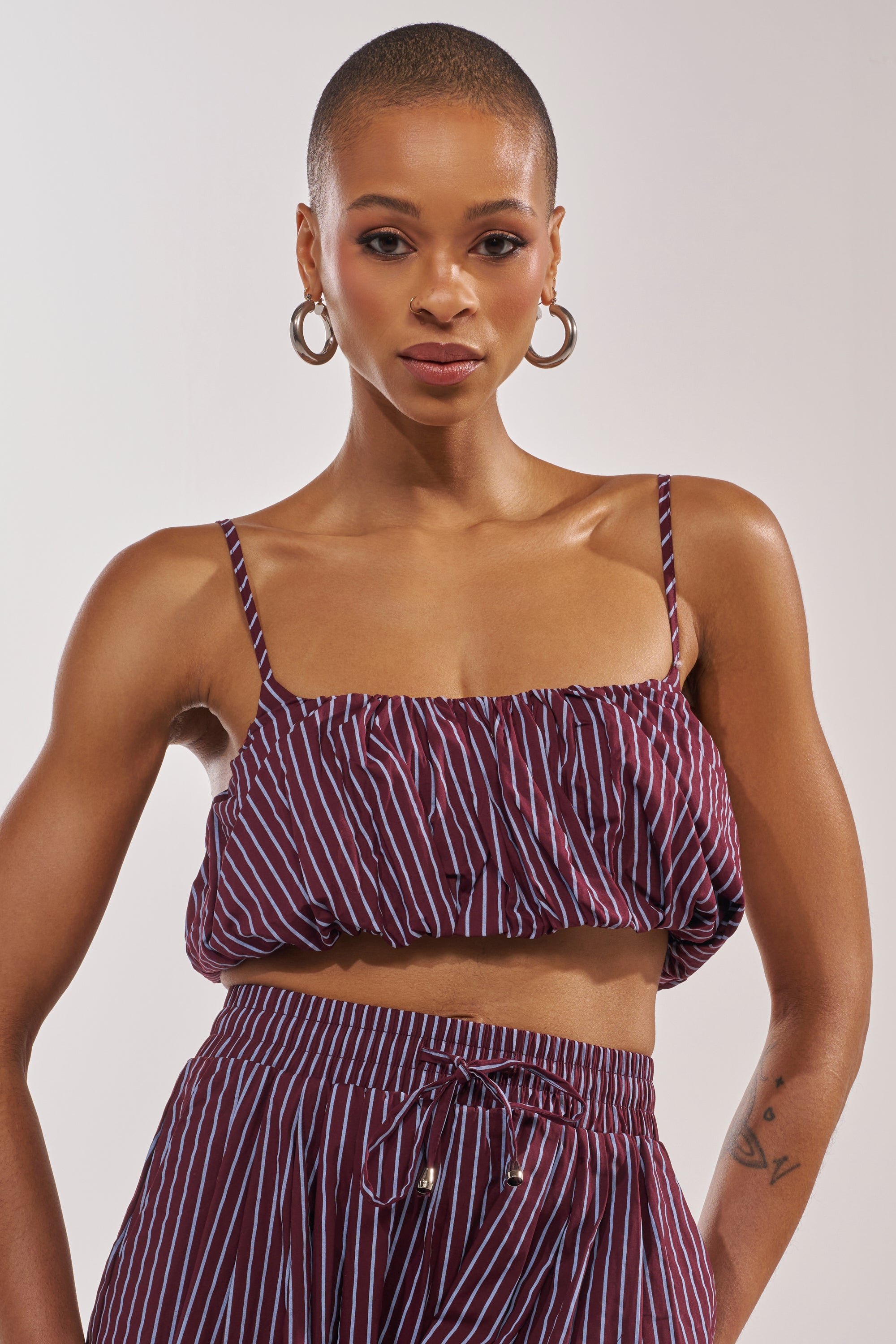 A woman with short hair wears large hoop earrings and the COULD BE DIFF BRALETTE in maroon and blue stripes, paired with matching high-waisted pants, as she stands confidently before a plain light background. 0