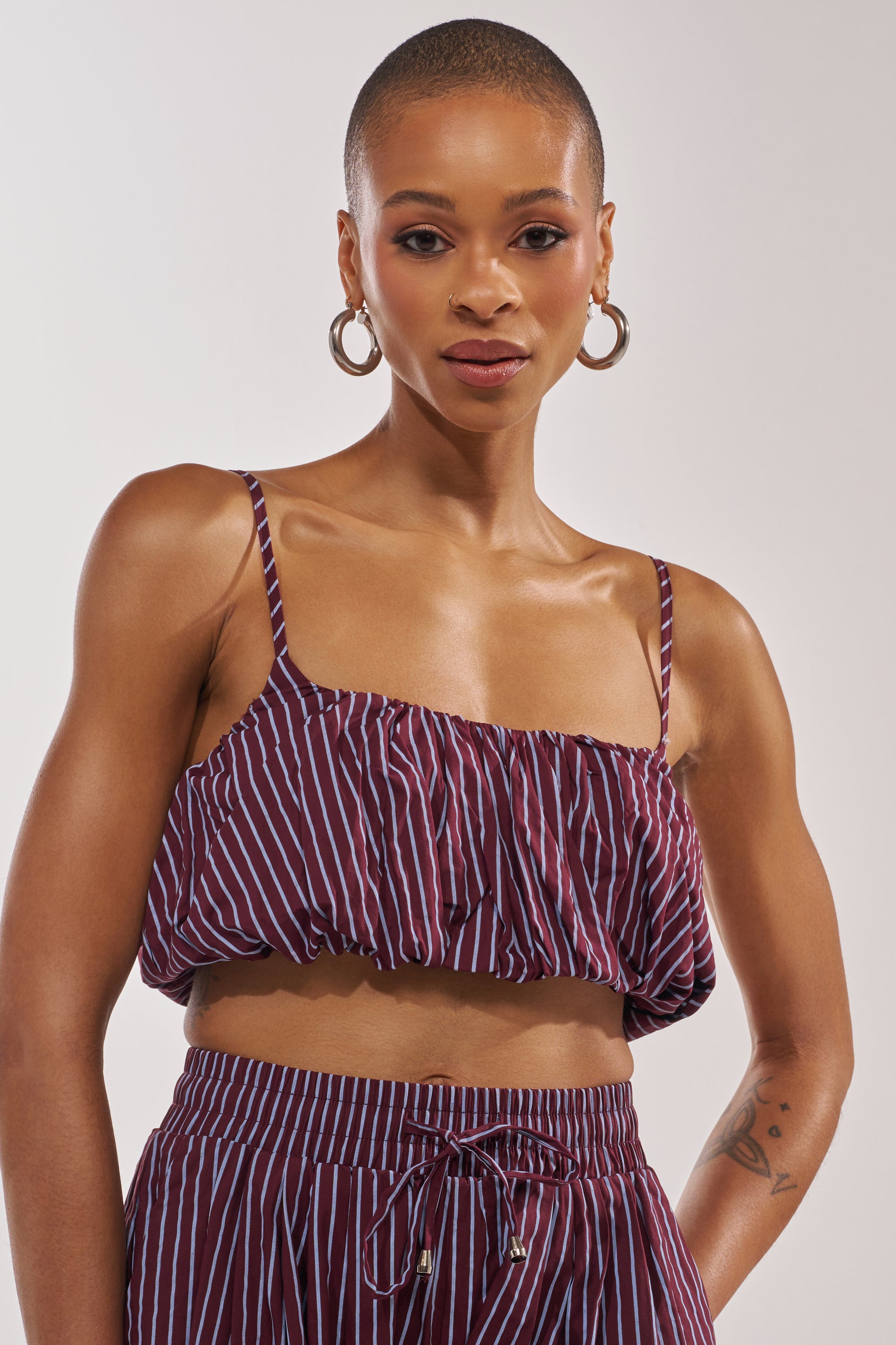 A woman with a shaved head and large hoop earrings wears the COULD BE DIFF BRALETTE in a maroon and blue stripe, paired with matching drawstring pants, posing confidently for a summer style against a neutral backdrop. 1