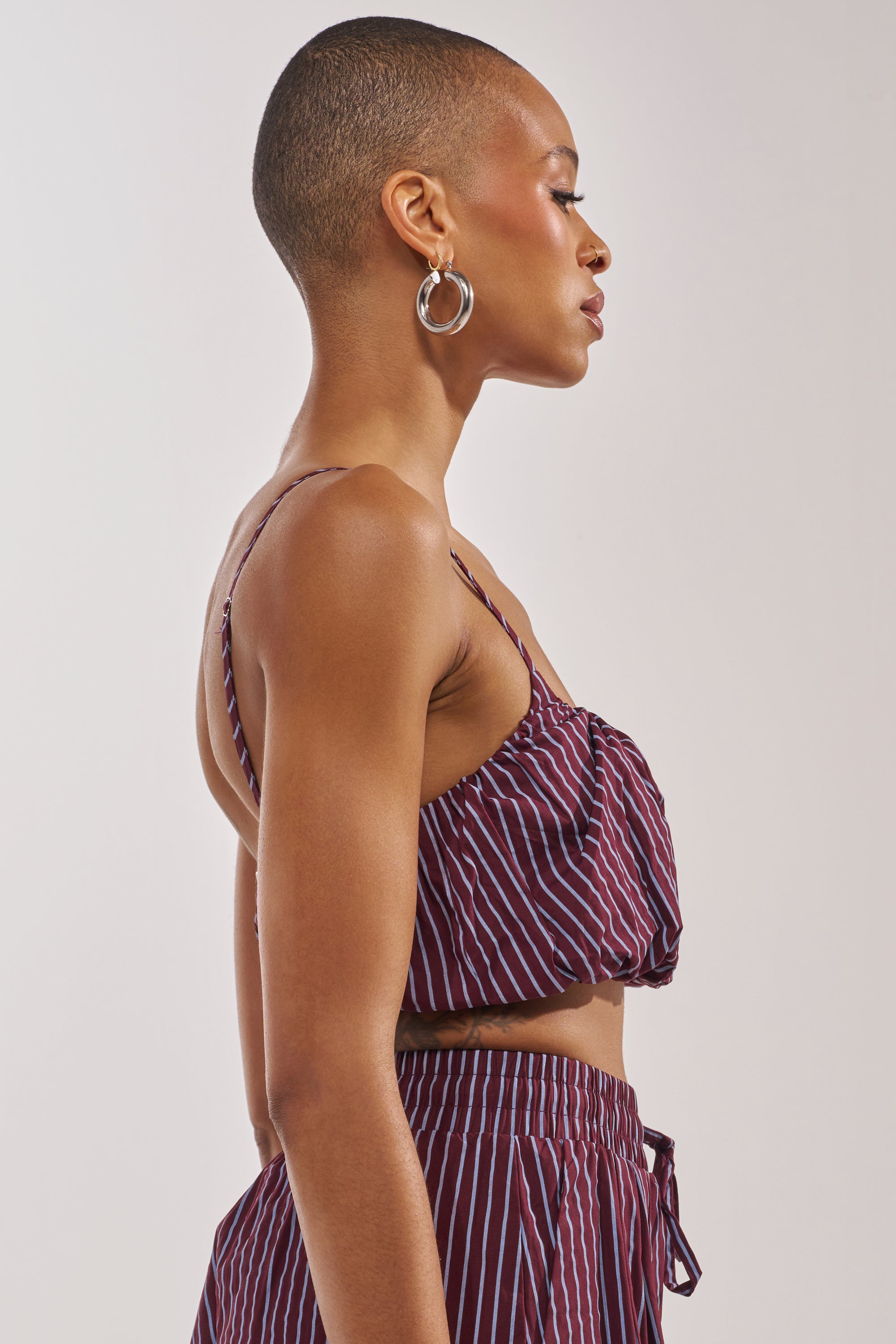 A woman with a shaved head wears silver hoop earrings and the COULD BE DIFF BRALETTE, a maroon and white striped festival top, with matching pants. She is in profile against a plain light background. 2