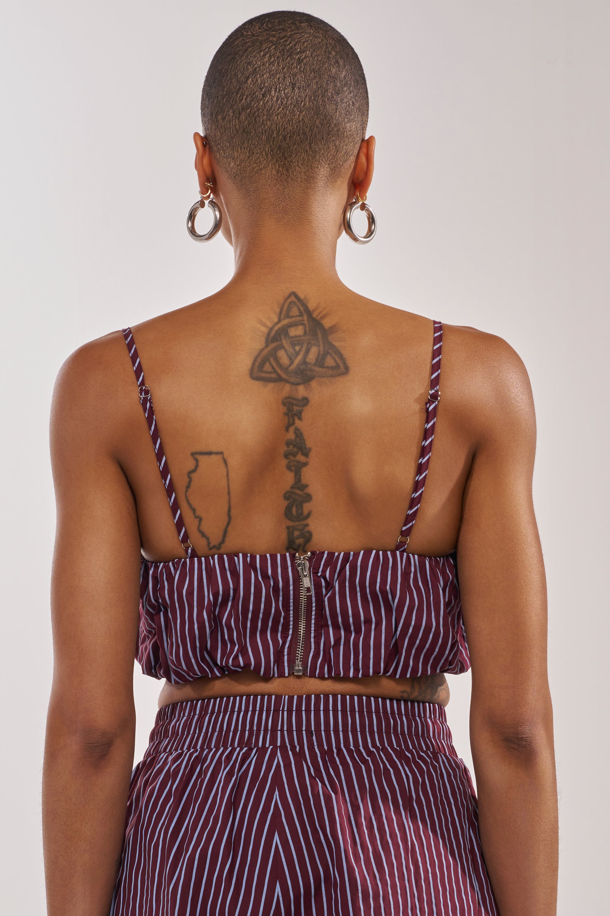 A person with a shaved head, large hoop earrings, and tattoos—including a triquetra, “PATCHEZ,” and the outline of Illinois—is shown from the back wearing the COULD BE DIFF BRALETTE in maroon. 3