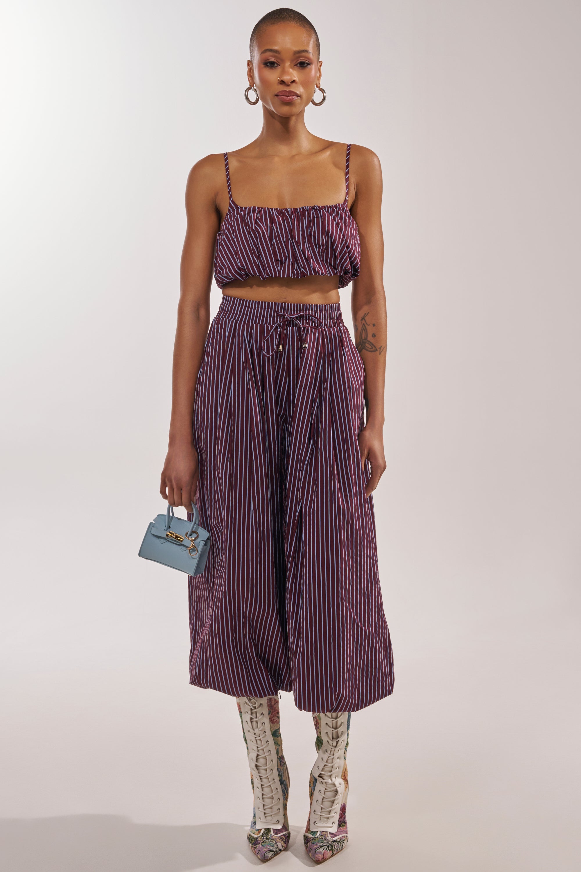 A woman wears the COULD BE DIFF BRALETTE, a maroon and blue striped top with matching wide-leg pants, styled with large hoop earrings, a small blue handbag, and knee-high open-toe snakeskin boots. 5