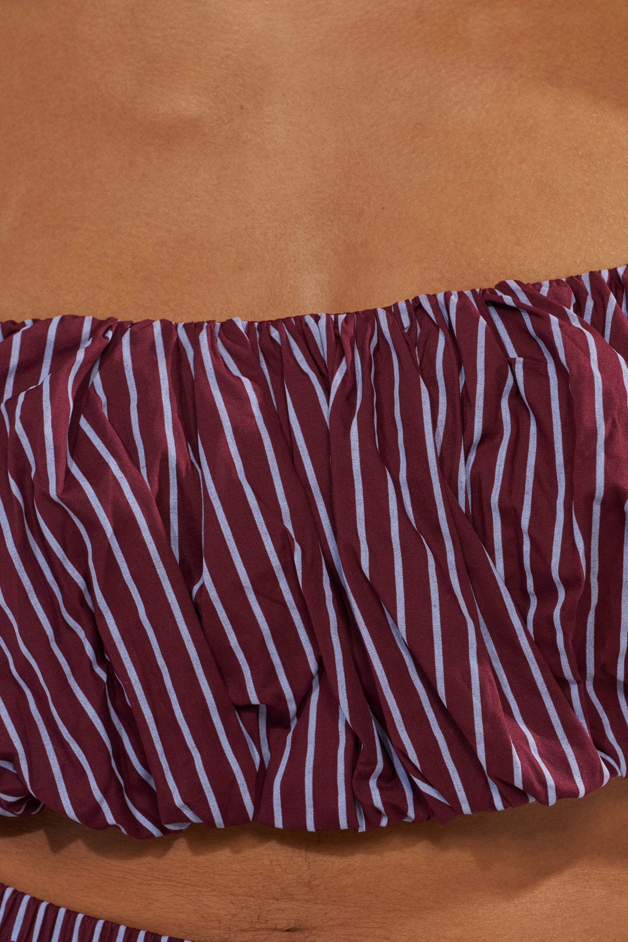 A close-up of a person wearing the COULD BE DIFF BRALETTE, featuring maroon fabric with white vertical and diagonal stripes, ruched texture, and tan skin—ideal as a trendy summer festival top. 7