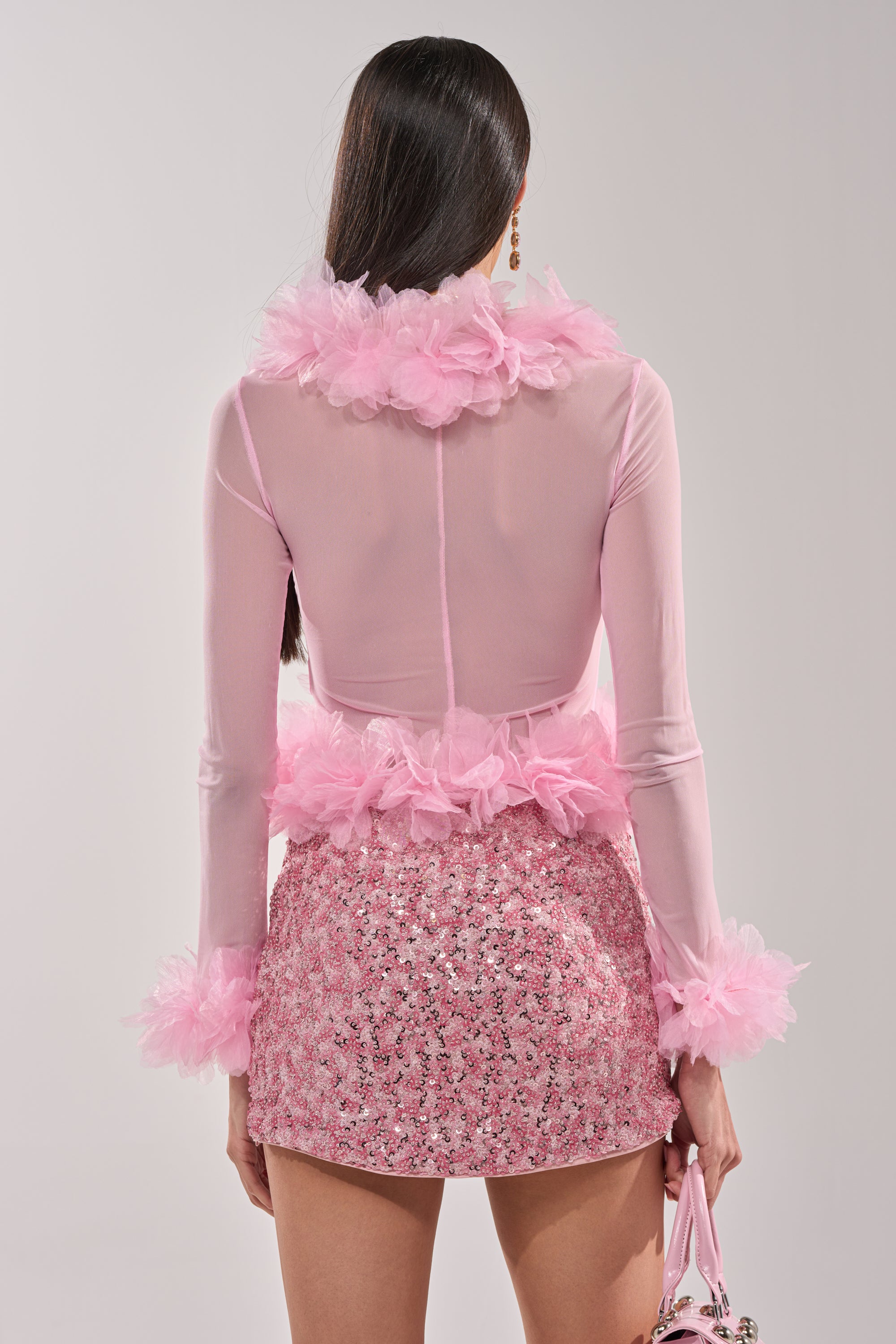 A woman with long, straight dark hair is seen from behind wearing the RUFFLE CITY LONG SLEEVE LIGHT PINK BLOUSE featuring sheer ruffles and tulle detailing, paired with a pink sequined mini skirt and holding a small handbag. 3