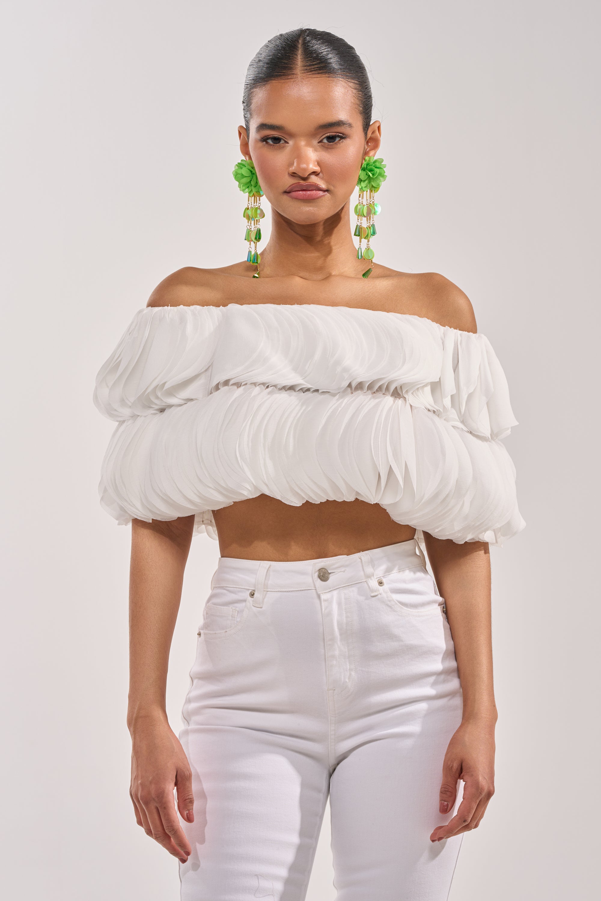 A woman wears the DAYDREAMER CHIFFON BLOUSE, a white off-shoulder layered top, paired with high-waisted white pants. Her slicked-back hair and bold green earrings complete the editorial-inspired look against a light background. 0