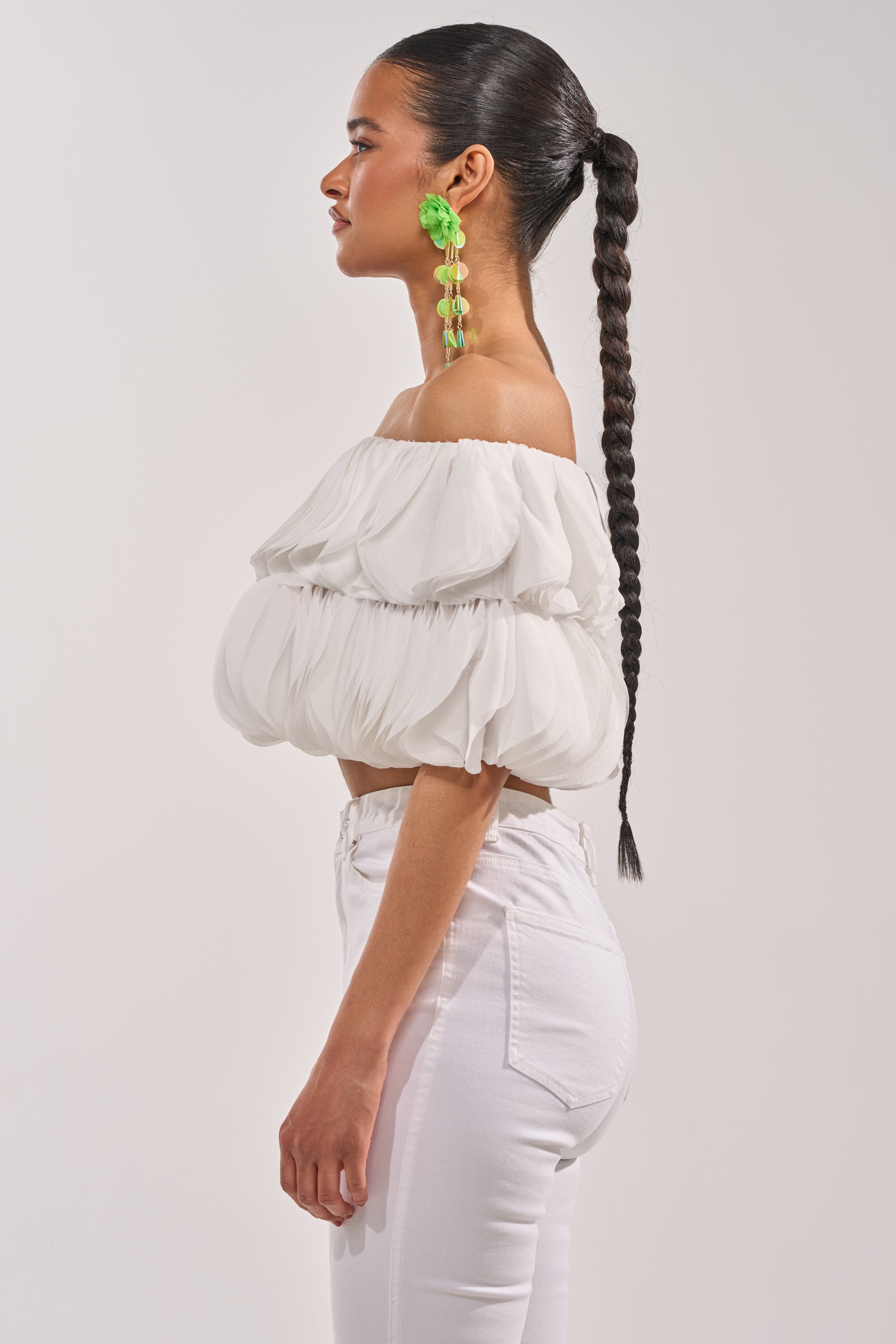 A woman in profile wears the DAYDREAMER CHIFFON BLOUSE—a white, high-fashion off-shoulder top with puff sleeves—paired with high-waisted white pants, bright green statement earrings, and a sleek braided ponytail. 2