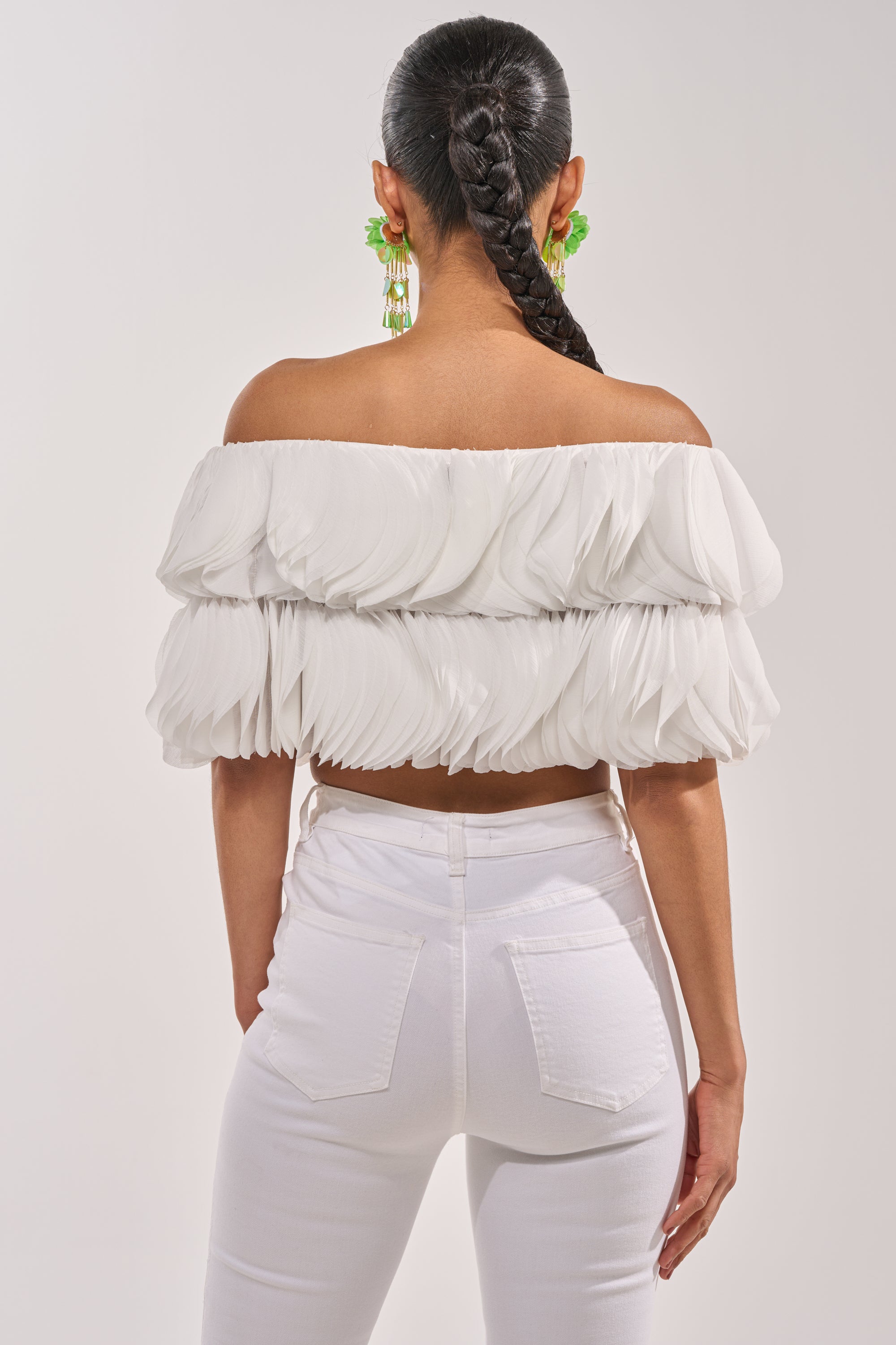 A woman with a long braided ponytail is seen from behind, wearing the DAYDREAMER CHIFFON BLOUSE off-shoulder style and white high-waisted jeans, accessorized with green statement earrings against a plain light background. 3