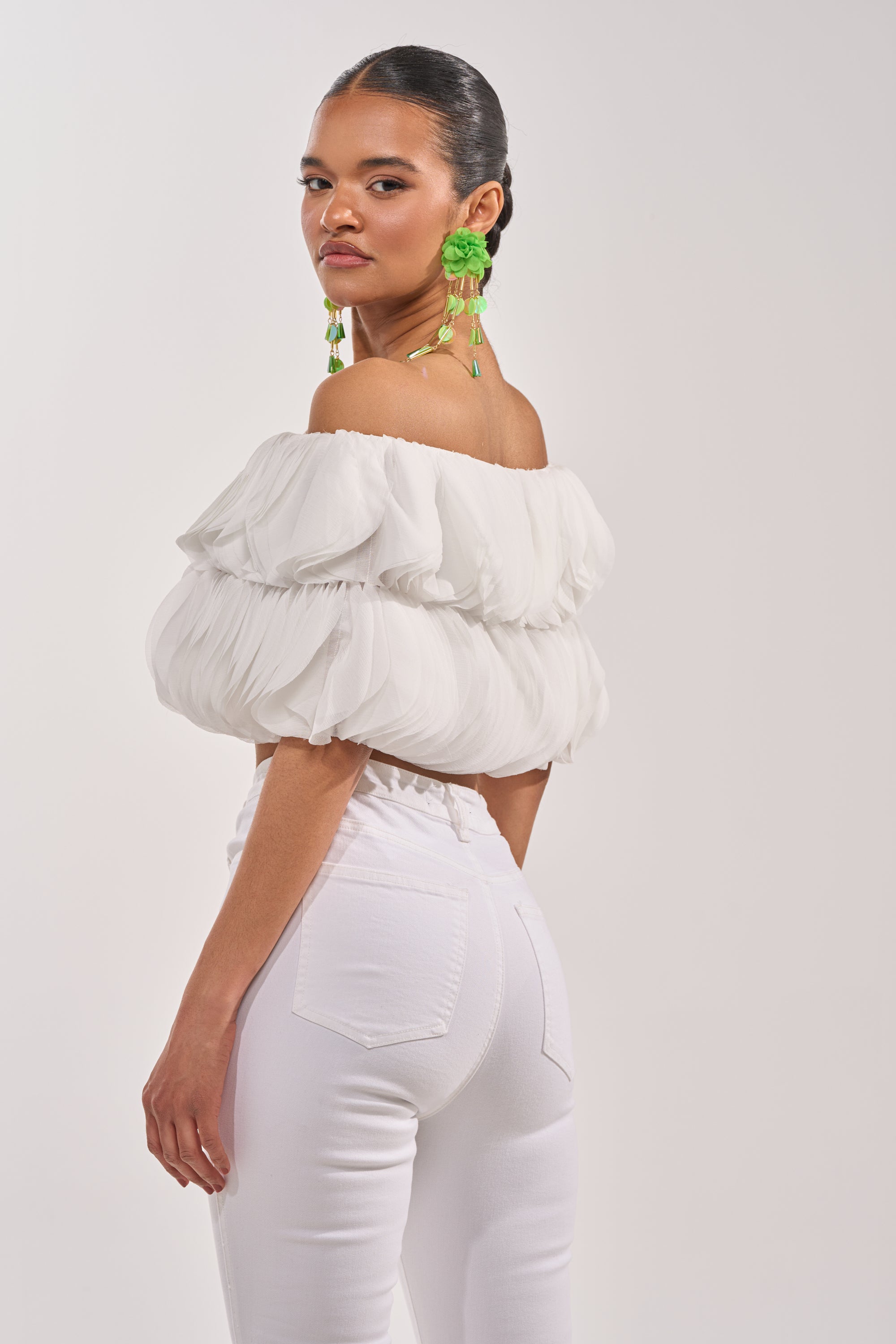 A woman poses confidently against a light background, wearing the DAYDREAMER CHIFFON BLOUSE—an off-shoulder white, ruffled top—paired with fitted white pants and bright green statement earrings. 4