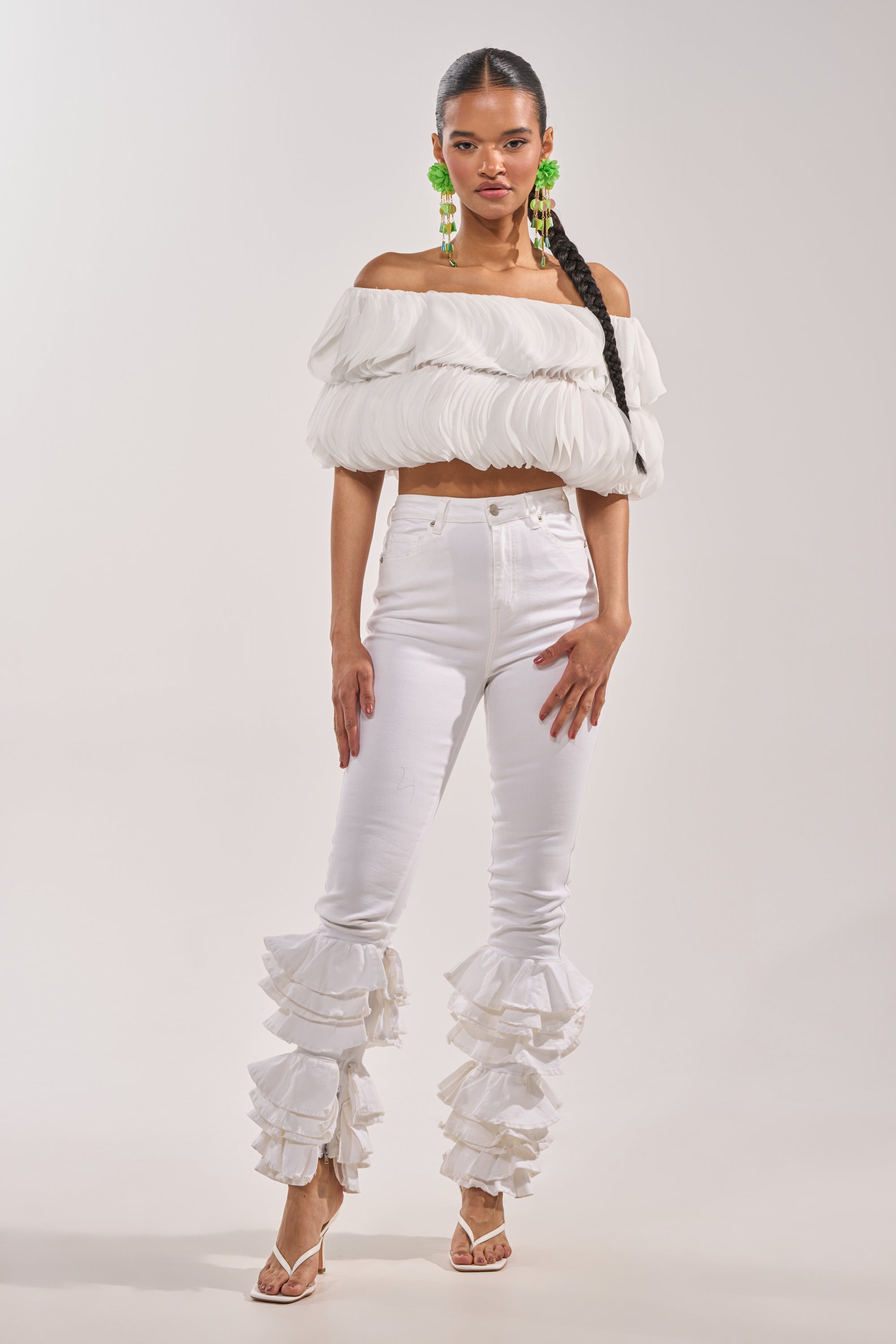A woman poses against a plain backdrop wearing the DAYDREAMER CHIFFON BLOUSE—an off-shoulder white top with ruffled details—paired with high-waisted ruffled white jeans, white sandals, bold green earrings, and a long braided hairstyle. 5