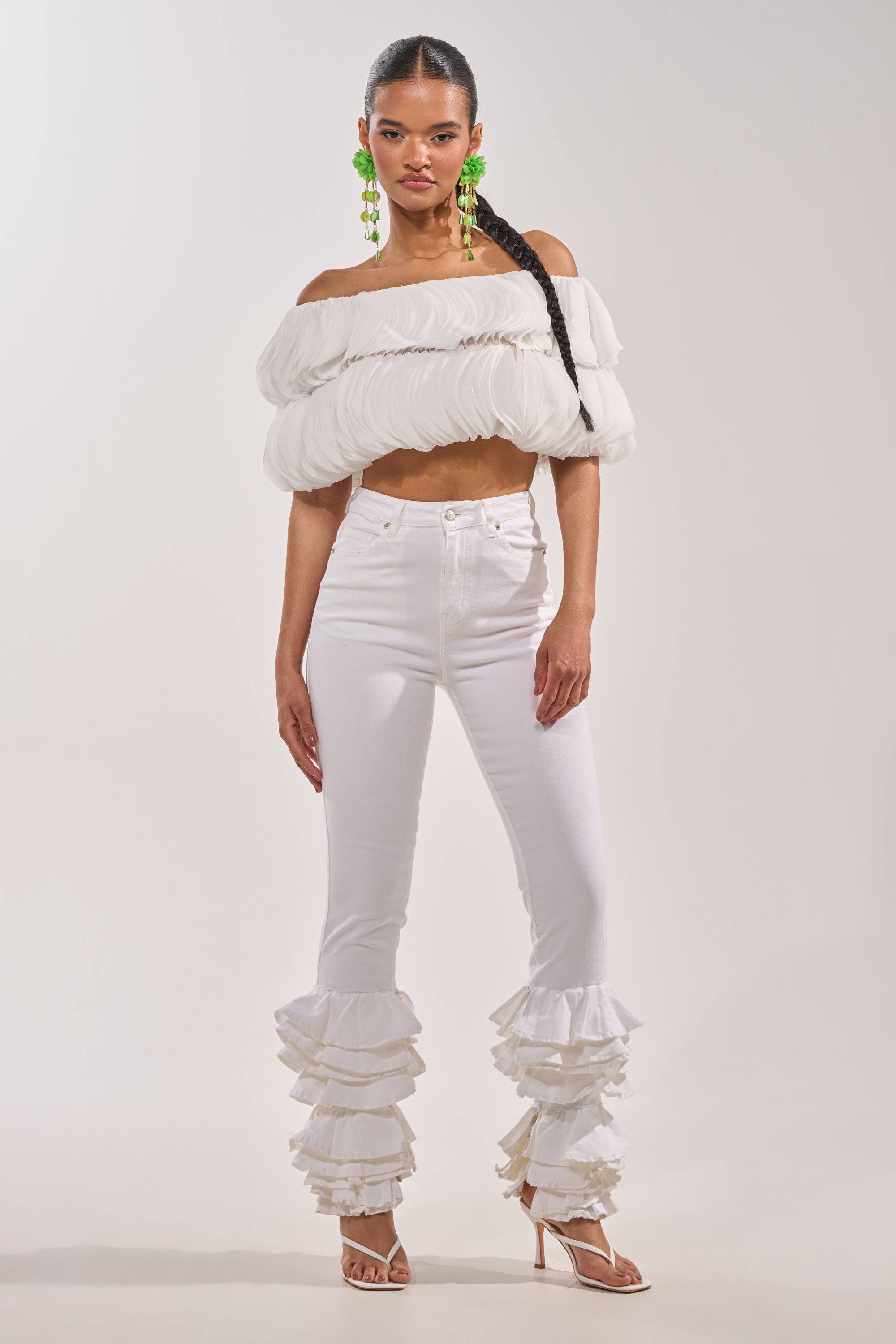 A woman poses against a plain background wearing the DAYDREAMER CHIFFON BLOUSE, paired with high-waisted white pants with ankle ruffles, strappy heels, and large green statement earrings. 6
