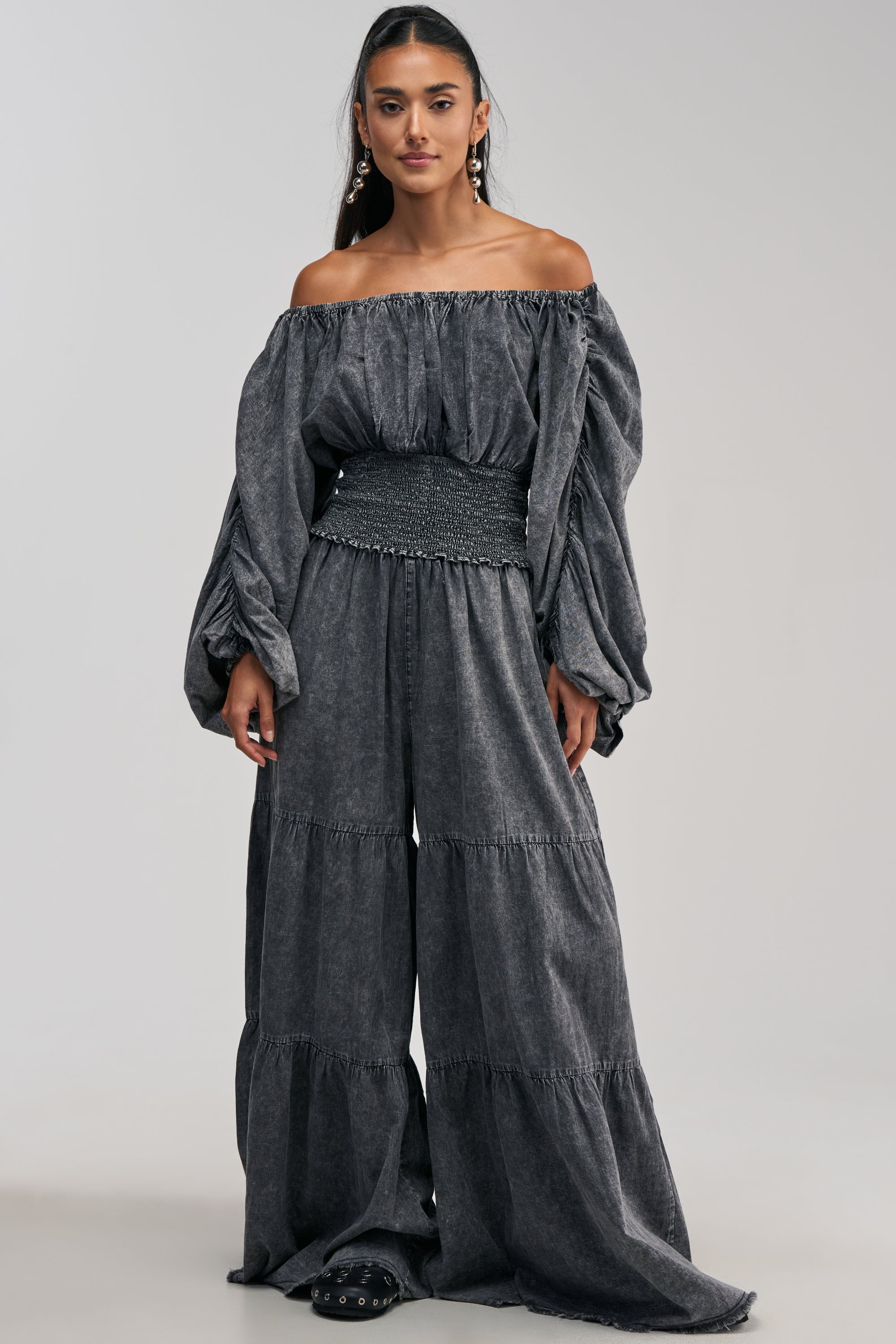A woman is wearing DENIM FOR DAYS PALAZZO PANT IN DARK DENIM, styled with black studded shoes, and standing against a plain light background.