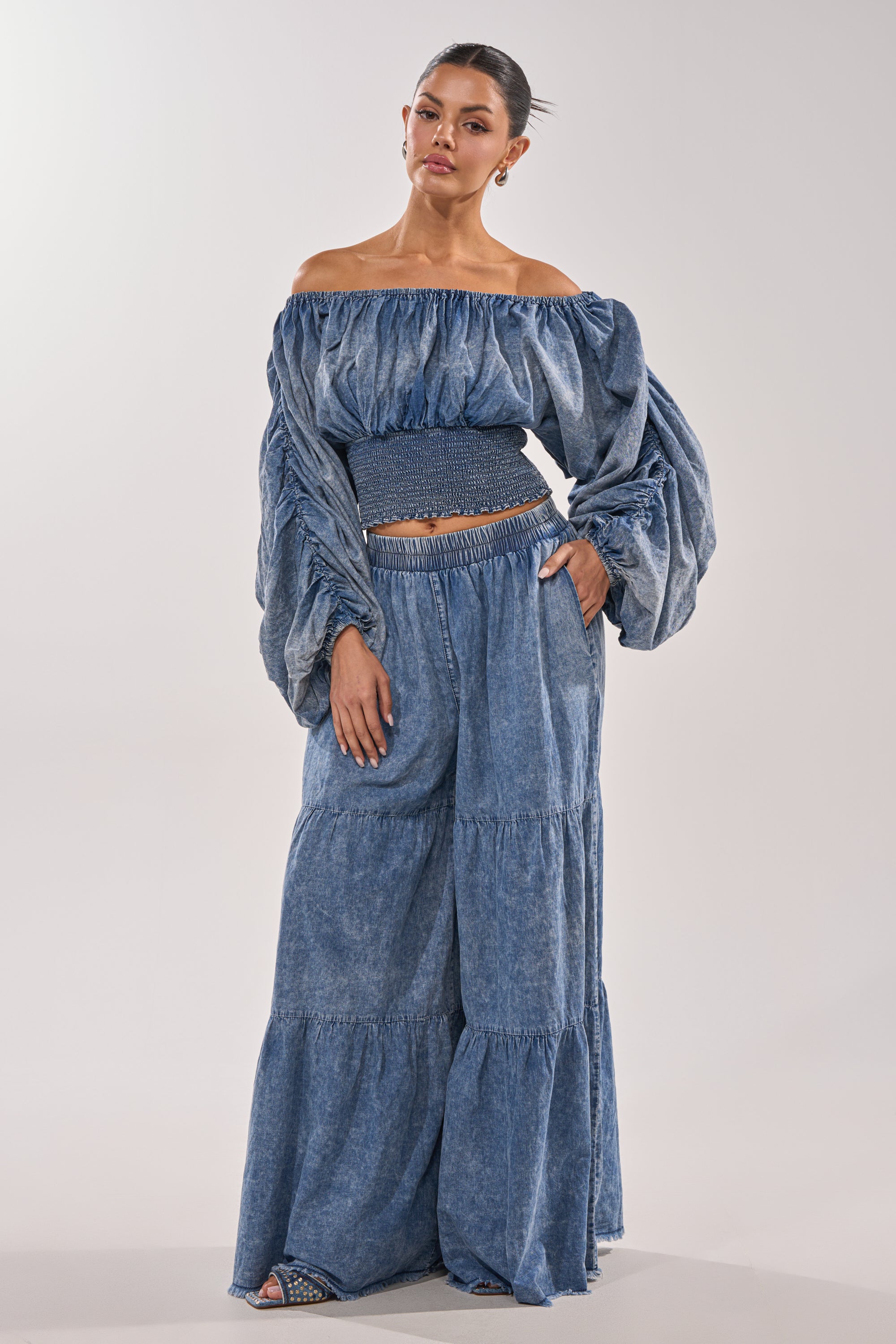 Wearing the DENIM FOR DAYS PALAZZO PANT, a woman styles the denim-hued, tiered palazzo pants with an off-the-shoulder voluminous-sleeved top, hoop earrings, and sandals, with her hair pulled back.