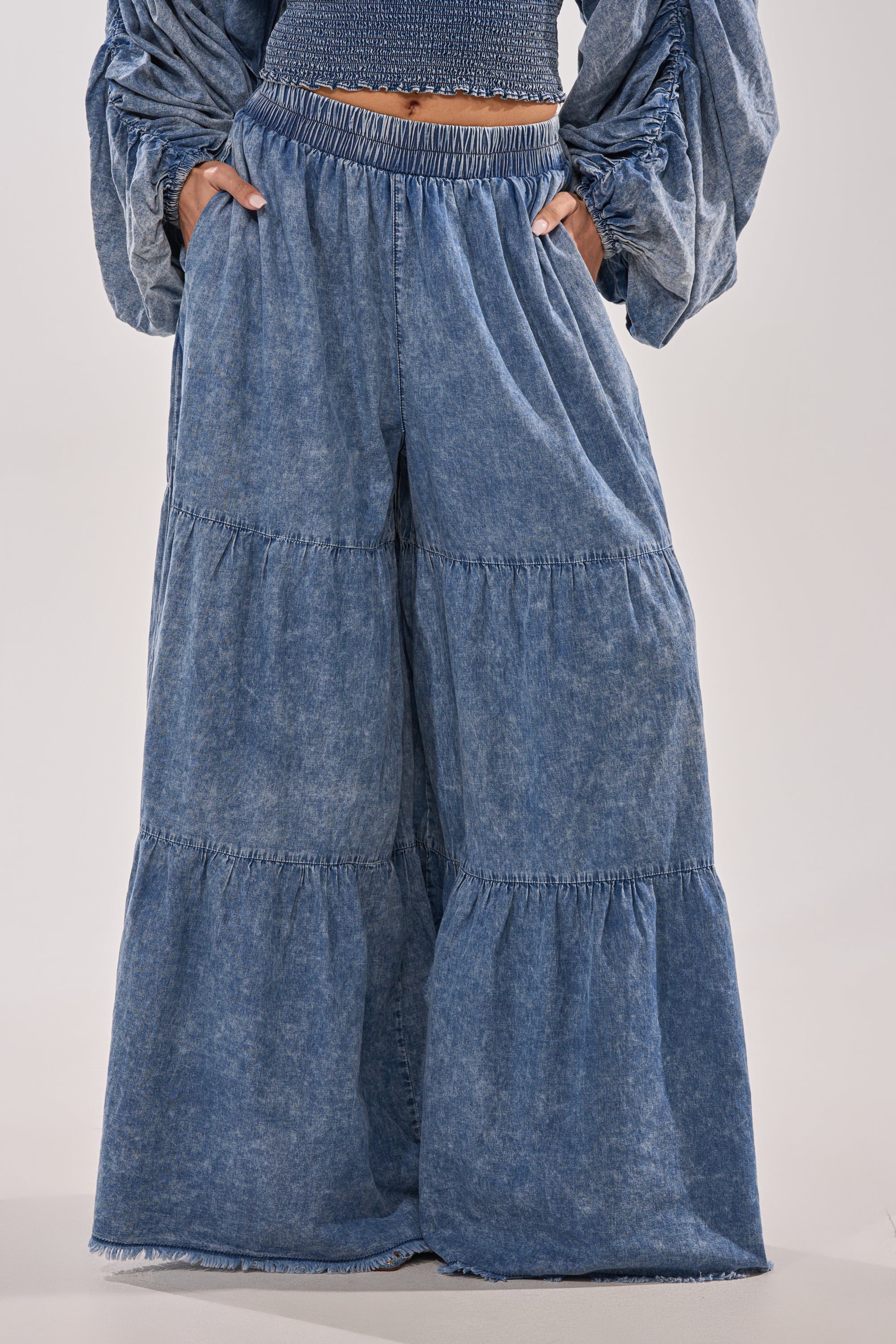 Wearing the DENIM FOR DAYS PALAZZO PANT with an elastic waistband and matching long-sleeve top, they channel relaxed boho style. Hands in pockets, the chest-down photo highlights the tiered denim design.