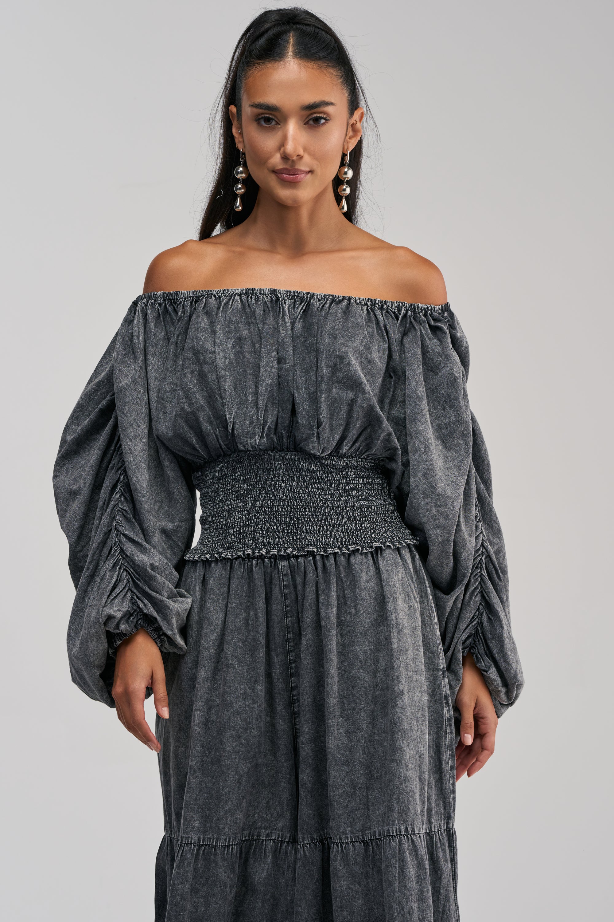 A woman models an off-the-shoulder, long-sleeve dark gray dress featuring a smocked waistband and ruched sleeves as she poses against a plain light background—her style echoing the chic appeal of the DENIM FOR DAYS OFF THE SHOULDER TOP IN DARK DENIM.