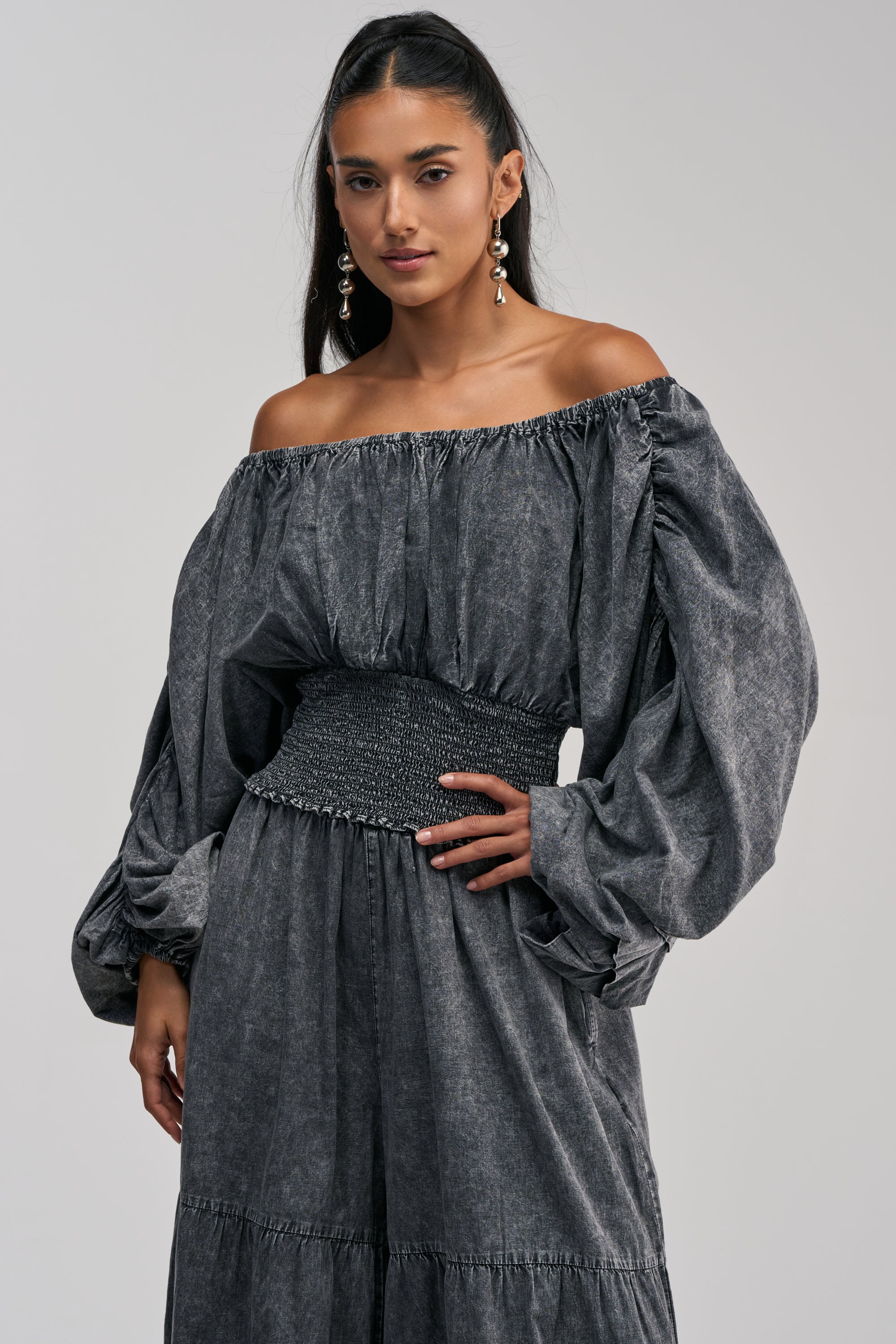 Model wearing the “Denim For Days Off The Shoulder Top in Dark Denim,” featuring voluminous sleeves and a smocked waist that echoes the casual vibe of denim. Styled with one hand on her hip against a plain background, she exudes effortless chic with her long dark hair and drop earrings.