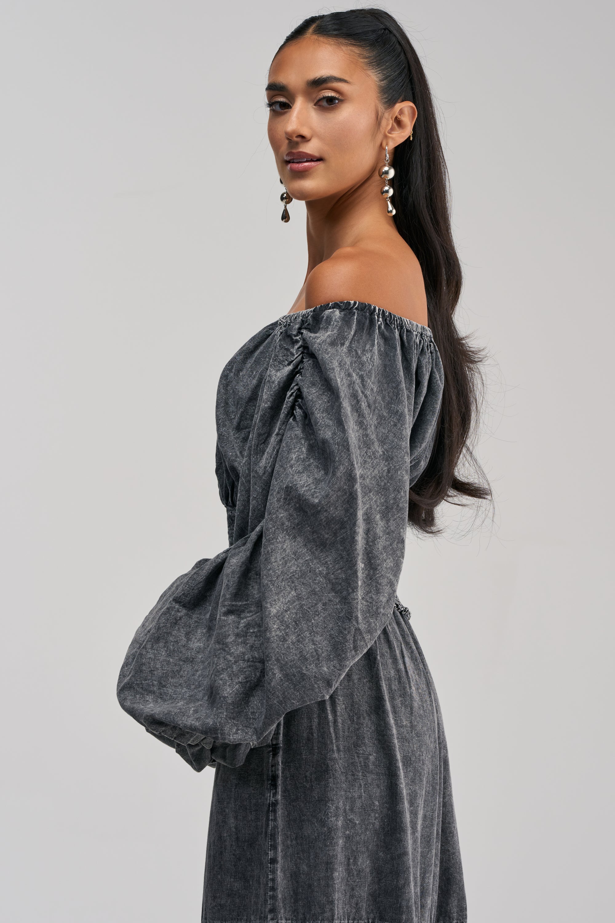 Woman with long dark hair in a high ponytail, wearing drop earrings and the DENIM FOR DAYS OFF THE SHOULDER TOP IN DARK DENIM, featuring long sleeves and a smocked waistband, stands sideways against a plain light background.