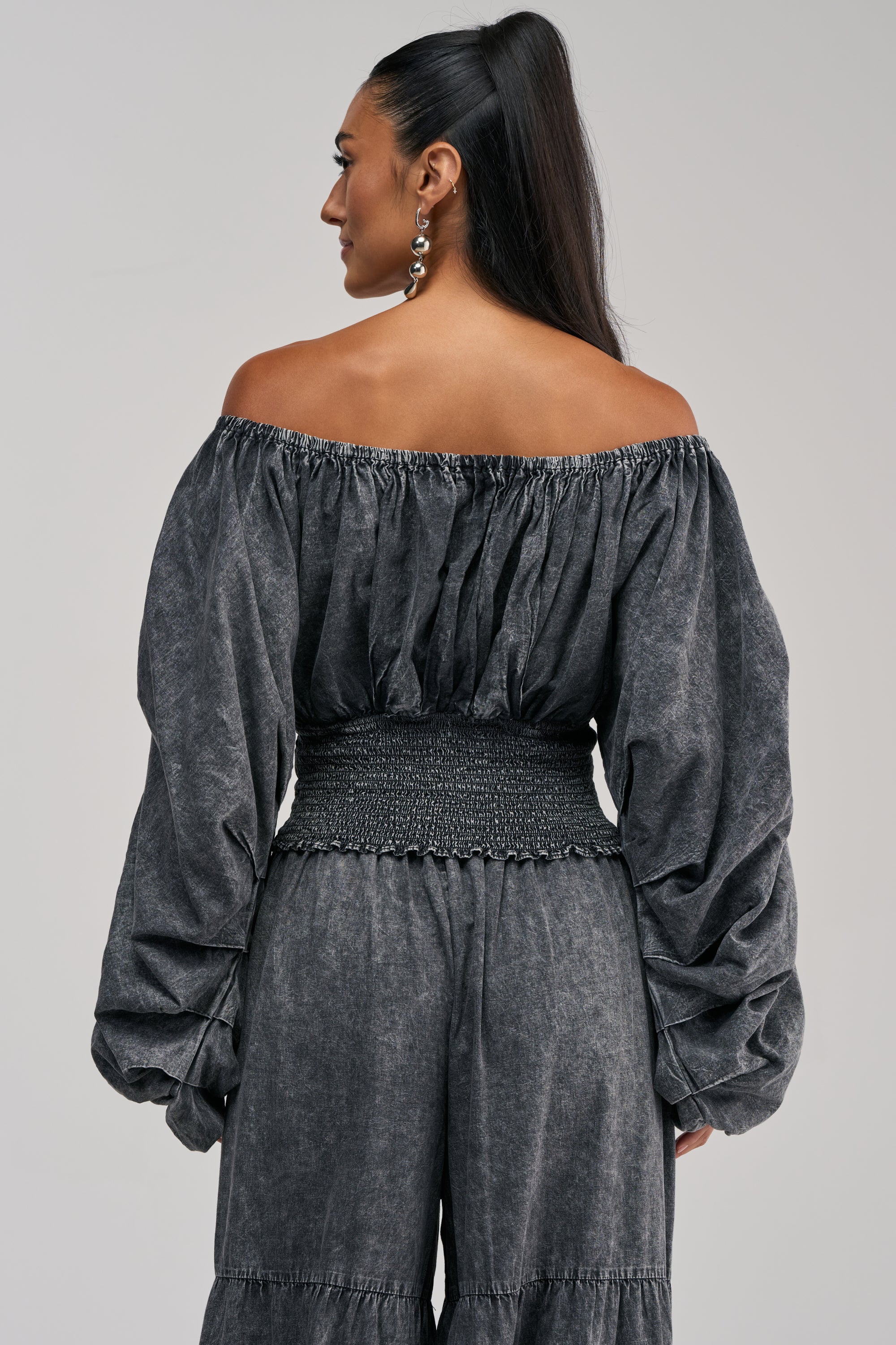 A woman is shown from behind wearing the DENIM FOR DAYS OFF THE SHOULDER TOP IN DARK DENIM, featuring long puffed sleeves and a smocked waist. She pairs it with matching high-waisted pants, styles her long dark hair in a ponytail, and accessorizes with dangle earrings.