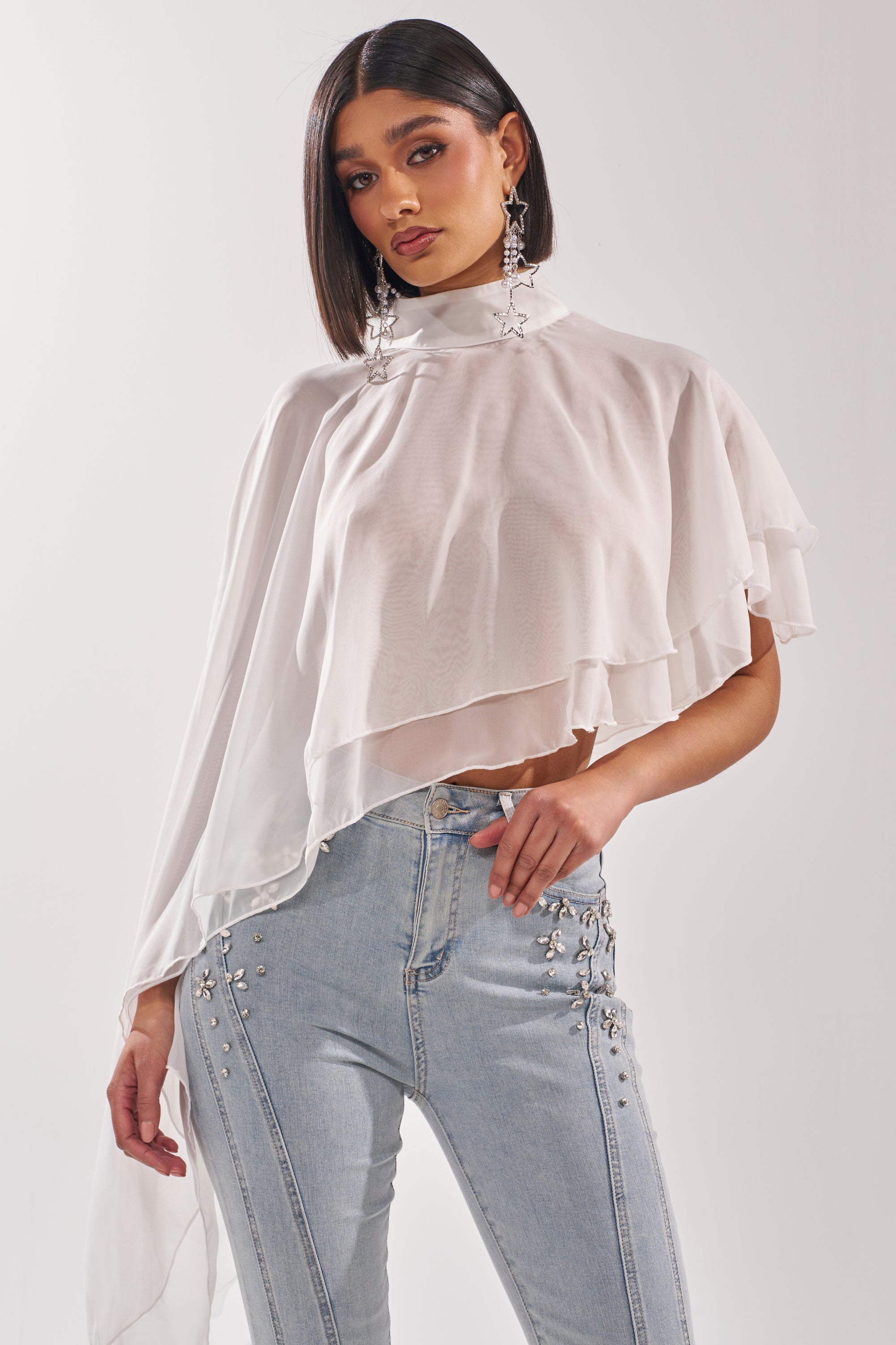 A woman wears the FOLLOW ME DRAPEY BLOUSE, a sheer white chiffon top with a mock neckline and asymmetrical flowy silhouette, paired with light blue high-waisted jeans. She has straight dark hair, star earrings, and stands against a white background. 1