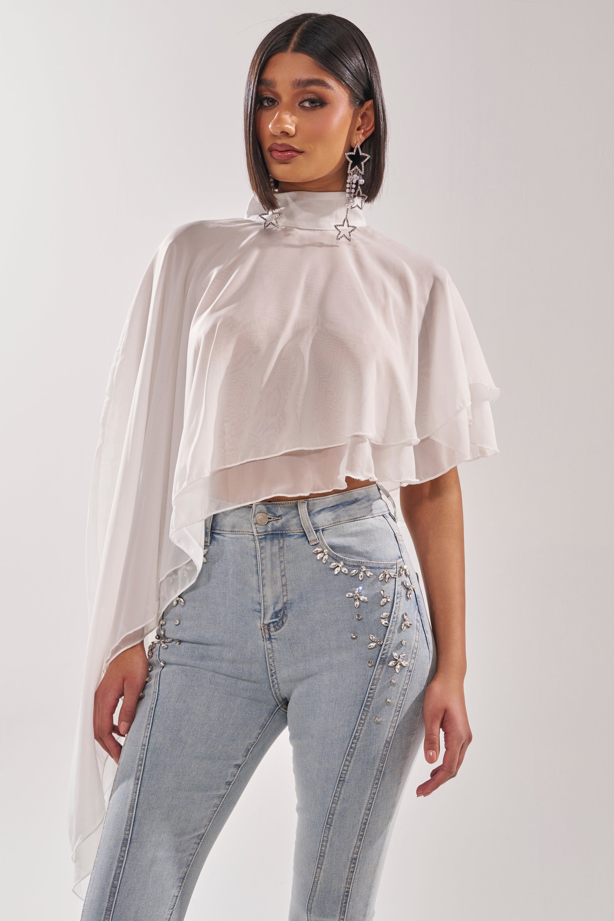 A woman wears the FOLLOW ME DRAPEY BLOUSE—white chiffon with an asymmetrical high mock neckline and layered, flowy fabric—paired with light blue, high-waisted embellished jeans against a plain background. 3