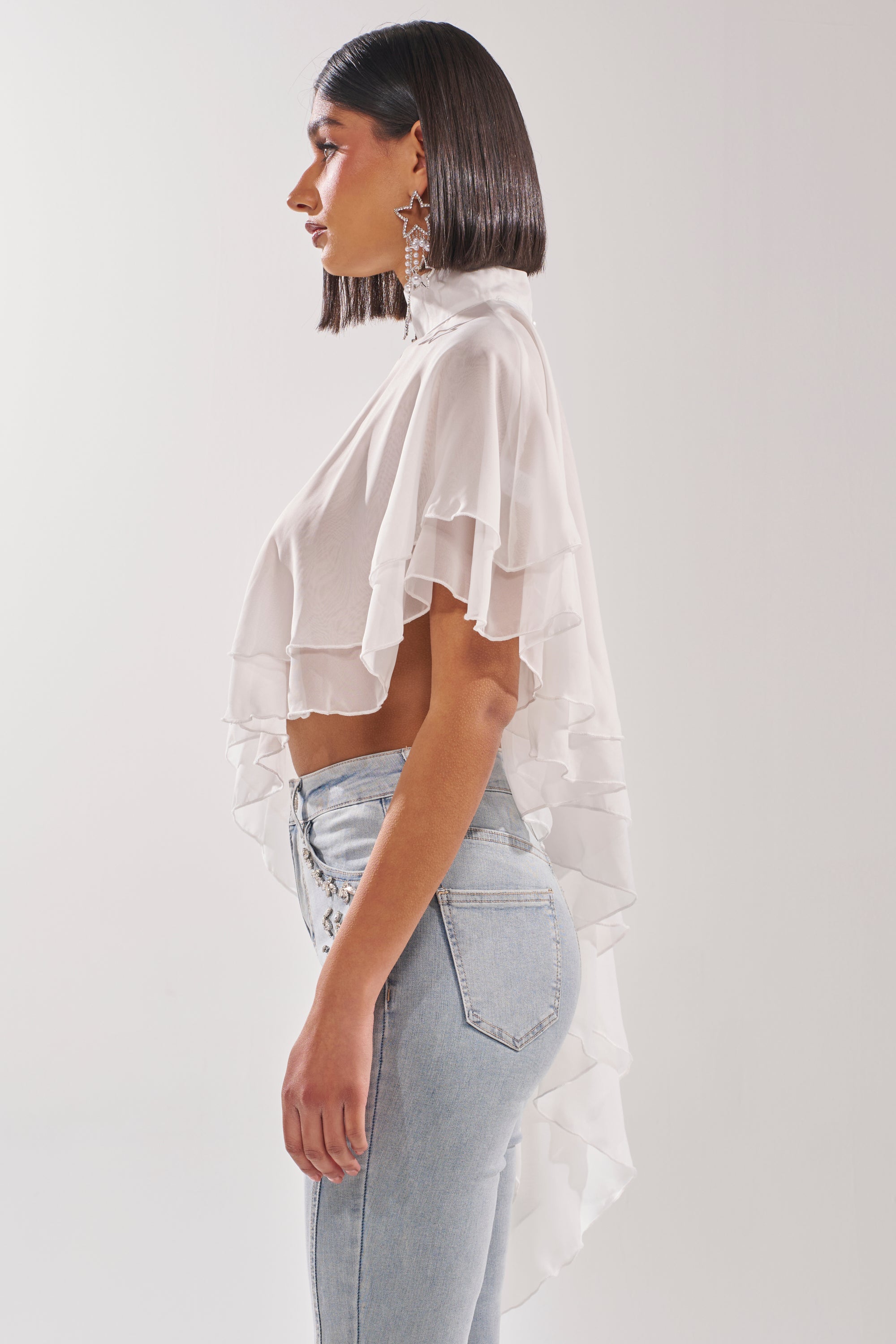A woman in profile wears the FOLLOW ME DRAPEY BLOUSE, a sheer, ruffled white chiffon top, paired with light-wash high-waisted jeans. She has straight shoulder-length dark hair and star-shaped earrings, posed against a plain white background. 4