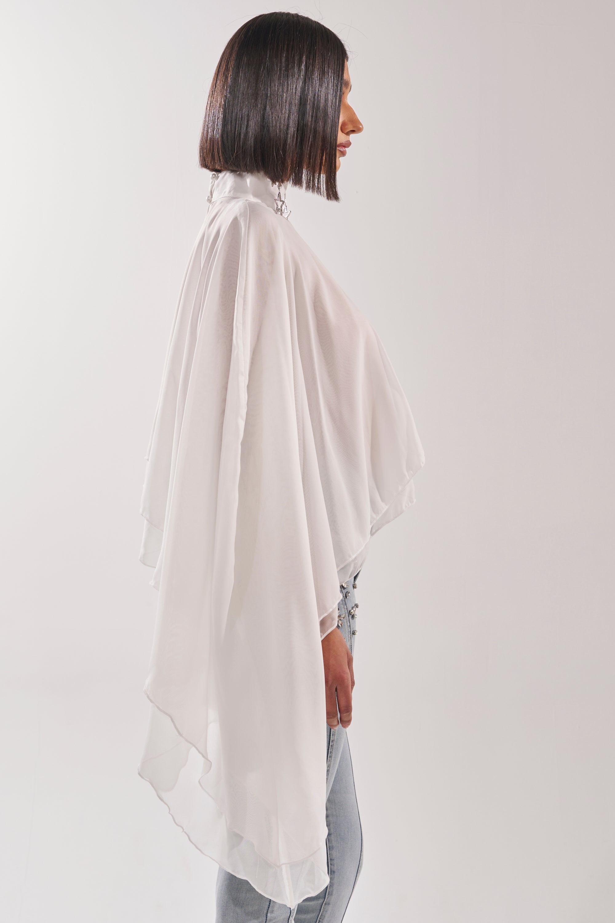 Wearing the FOLLOW ME DRAPEY BLOUSE, a person with straight, shoulder-length dark hair poses in profile, pairing the white chiffon top with light blue jeans against a plain light background. 5