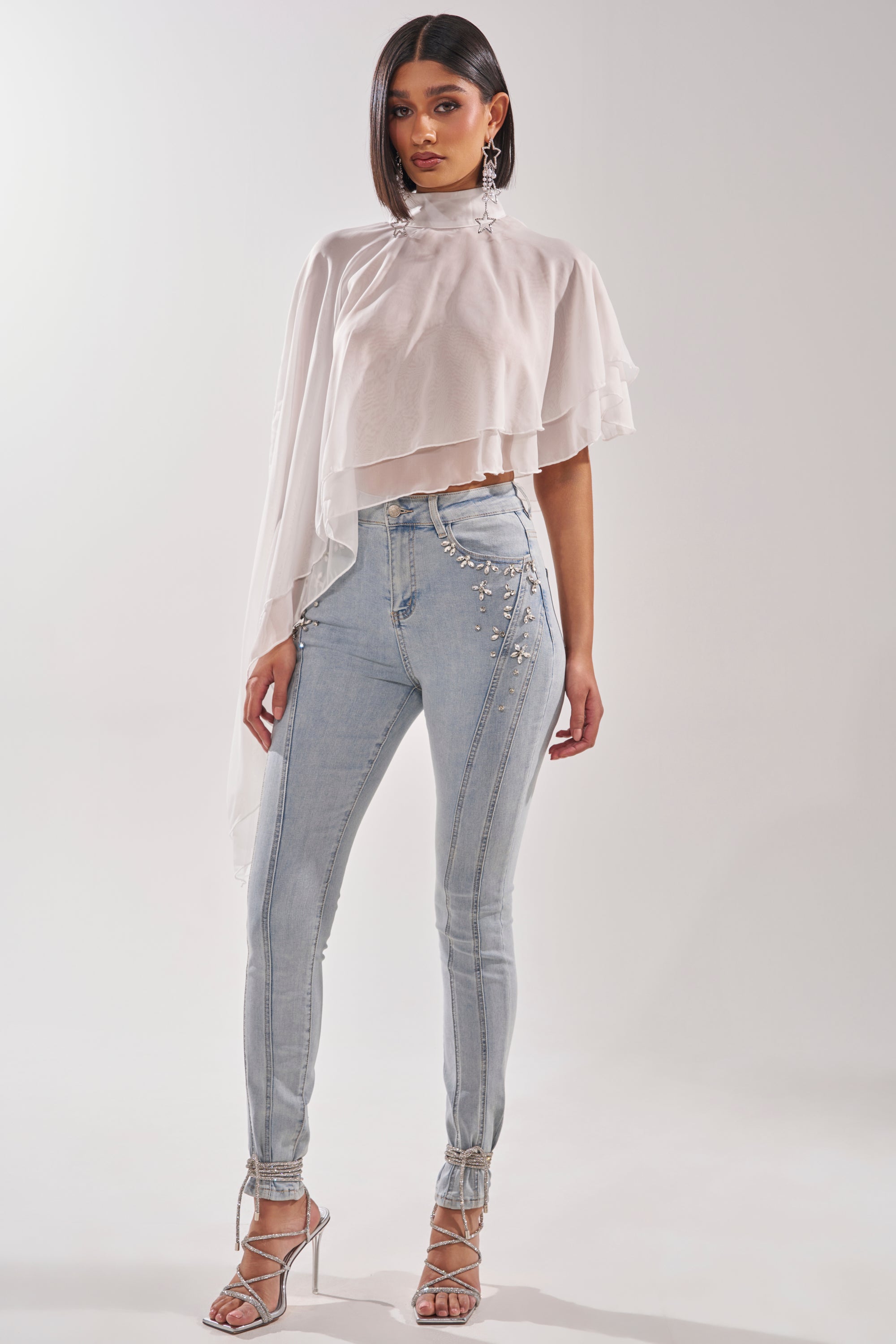 A woman with straight dark hair and a confident pose wears the FOLLOW ME DRAPEY BLOUSE— a sheer white chiffon top with an asymmetrical hem—paired with light-wash embellished skinny jeans, silver strappy heels, and statement earrings. 7