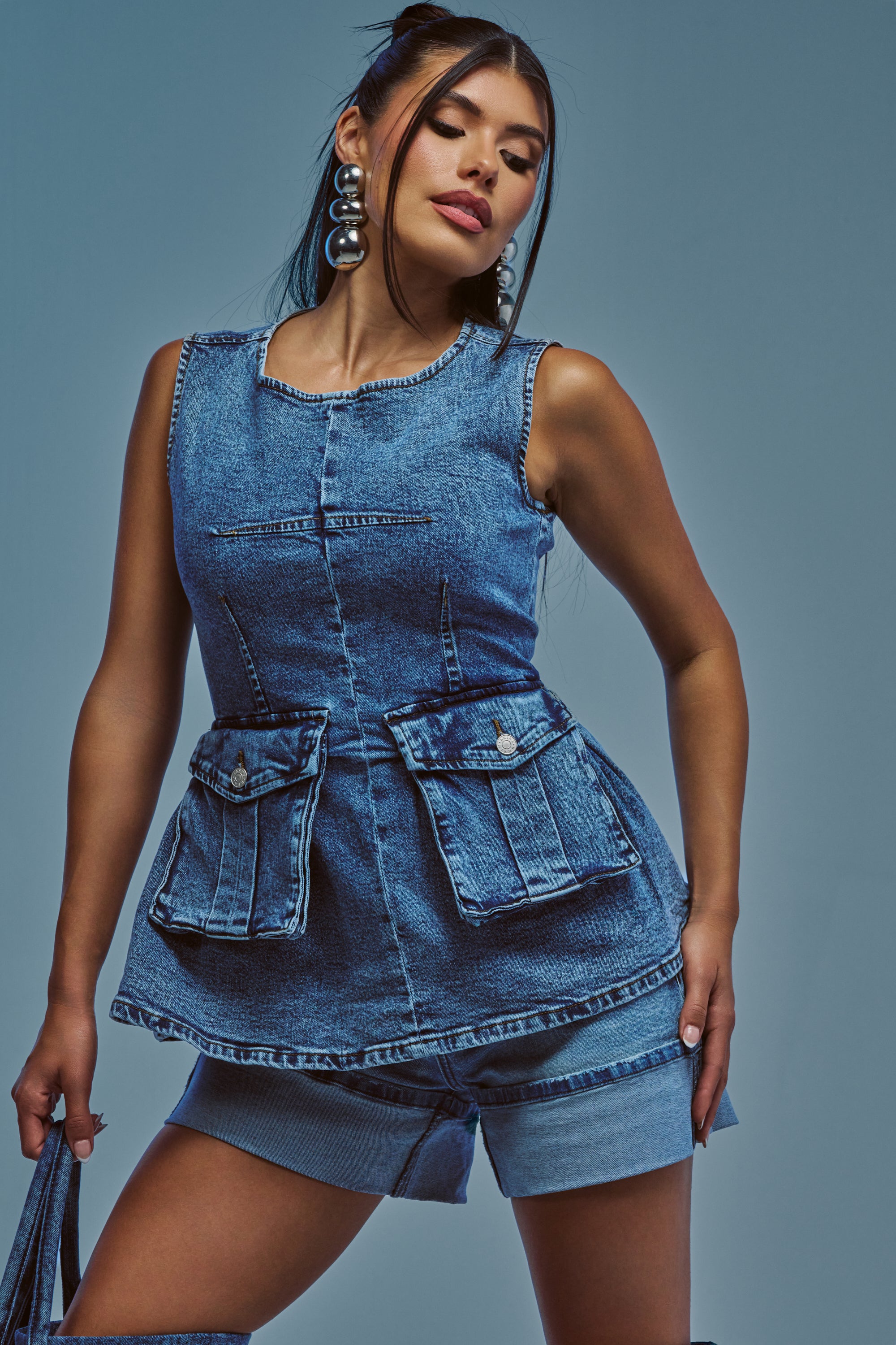 A woman poses against a blue background, wearing the TURN AROUND BLOUSE IN MEDIUM BLUE DENIM—a sleeveless, fitted denim peplum top with prominent front pockets—paired with matching shorts. She accessorizes with a bag in one hand and bold silver hoops, making an effortless summer streetwear statement.