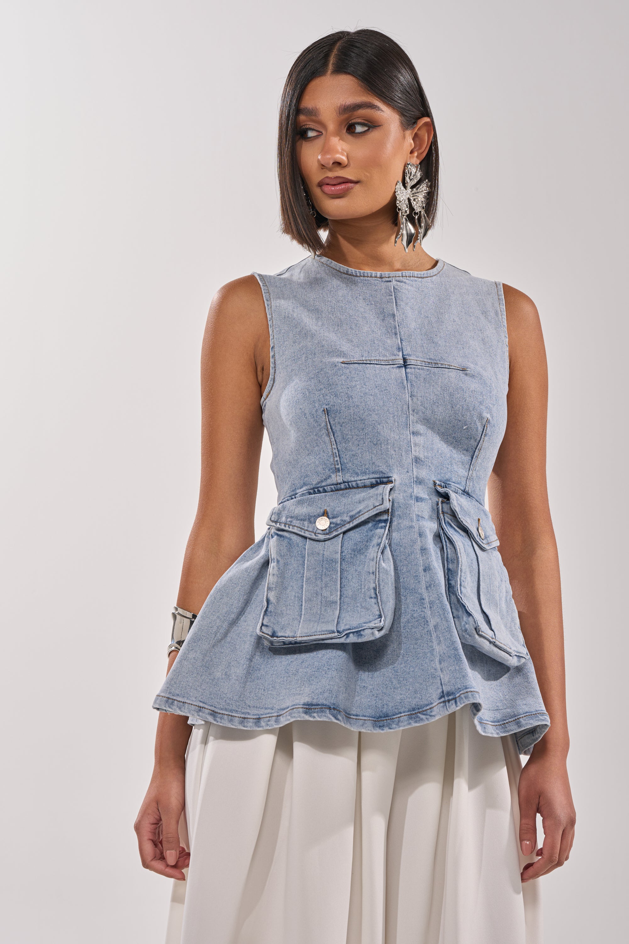 A woman with straight, shoulder-length dark hair wears the TURN AROUND DENIM BLOUSE as a sleeveless utility-chic top paired with white wide-leg pants, accessorized with statement earrings and a bracelet, standing against a plain background. 1