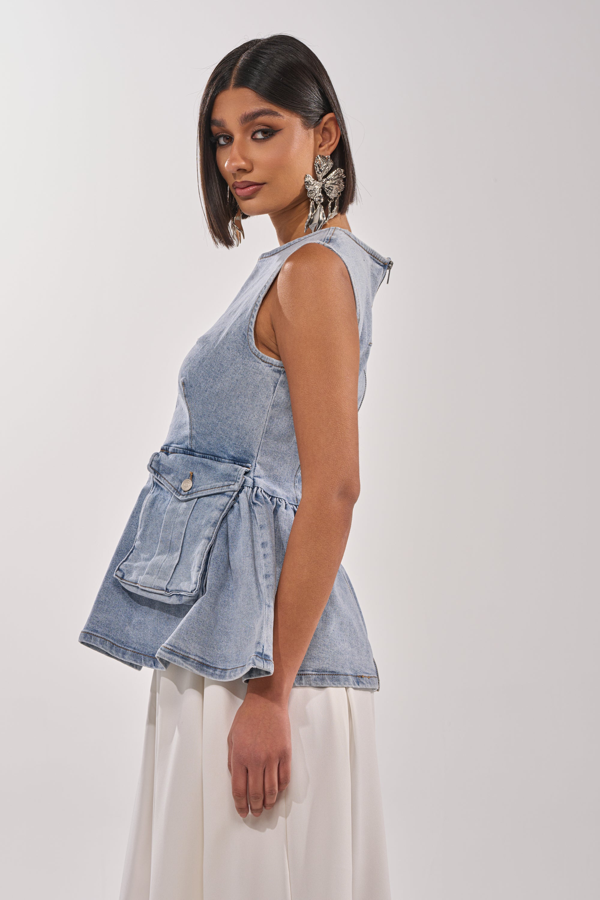 A woman with straight, shoulder-length dark hair wears statement earrings and the TURN AROUND DENIM BLOUSE—a sleeveless peplum top with a front pocket—paired with a white skirt, standing against a plain background. 2