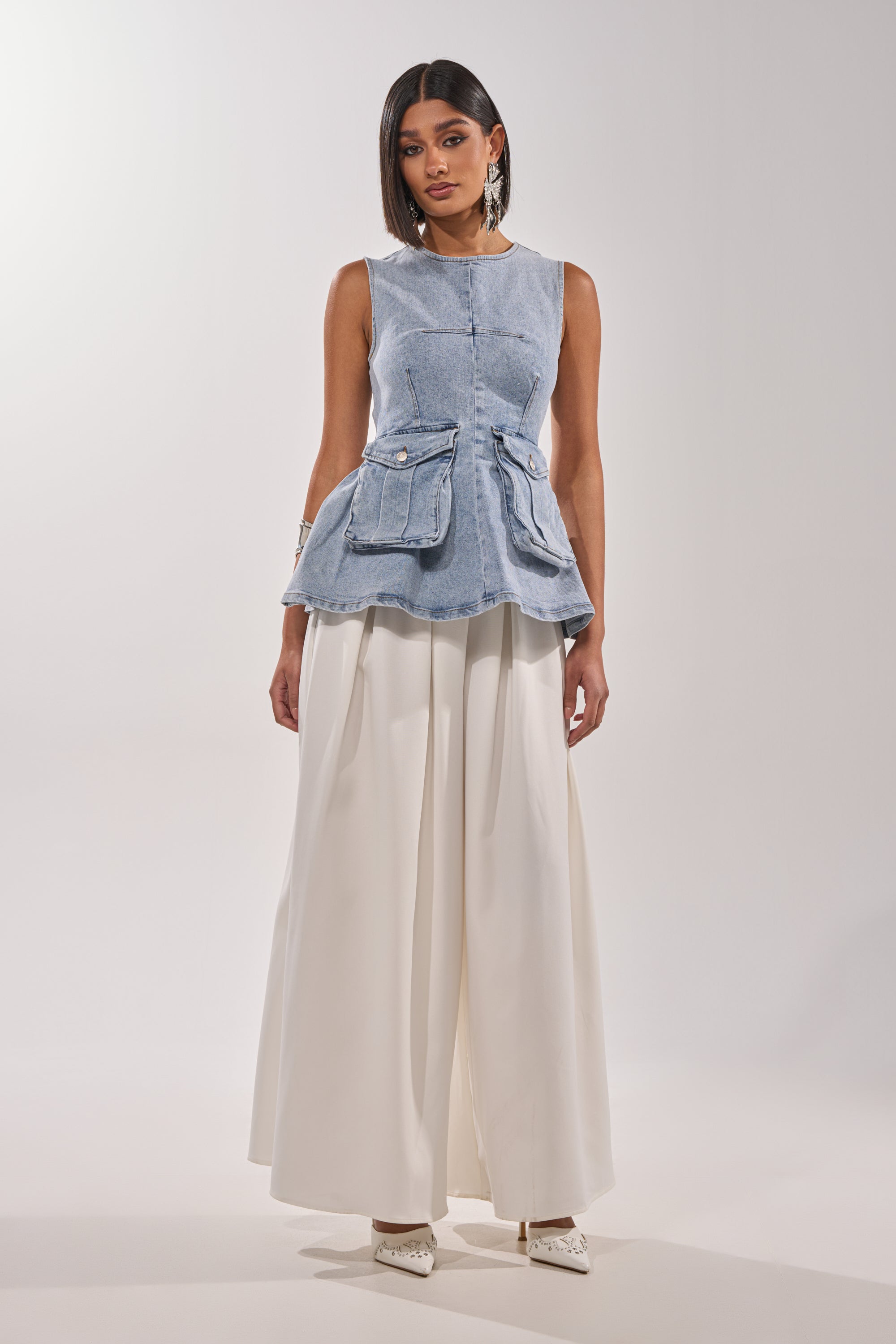 A woman stands against a plain background wearing the TURN AROUND DENIM BLOUSE in a utility-chic, light-wash style with front pockets, paired with wide-legged, high-waisted white trousers and white pointed shoes. 4