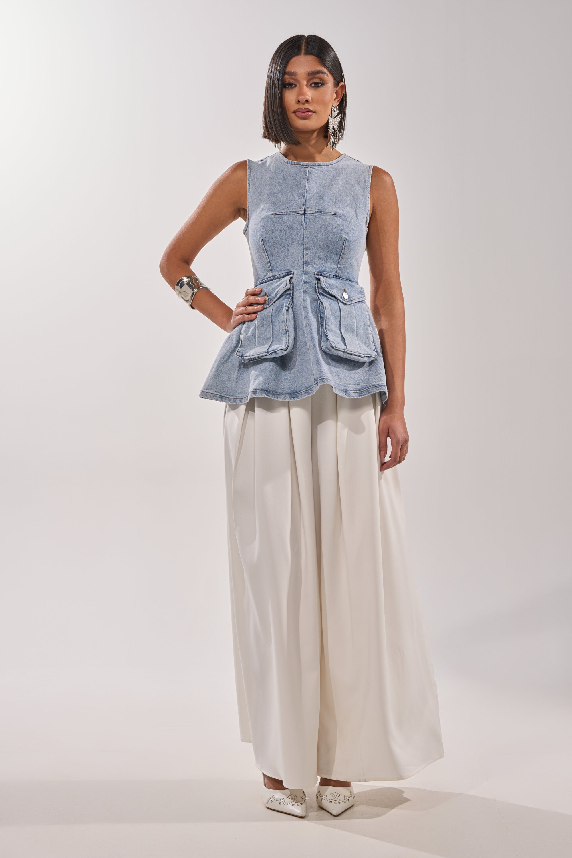 A woman stands against a plain background wearing the TURN AROUND DENIM BLOUSE, styled with wide-leg white trousers, white pointed shoes, and bold silver jewelry for an effortless utility-chic look. 5